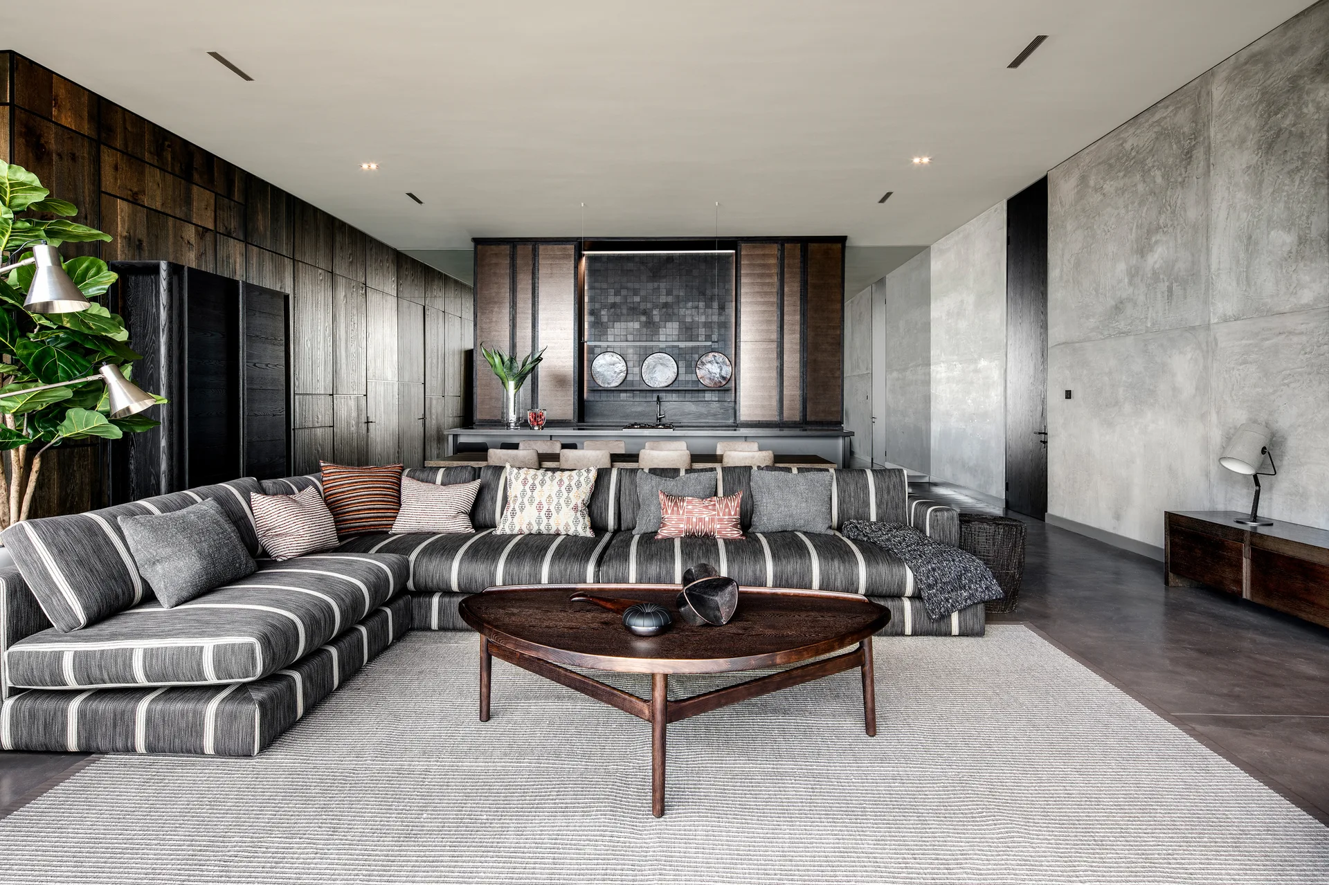 A large striped sectional sofa sits on a textured rug in an open-plan living room with a dark wood paneled wall and a concrete wall.