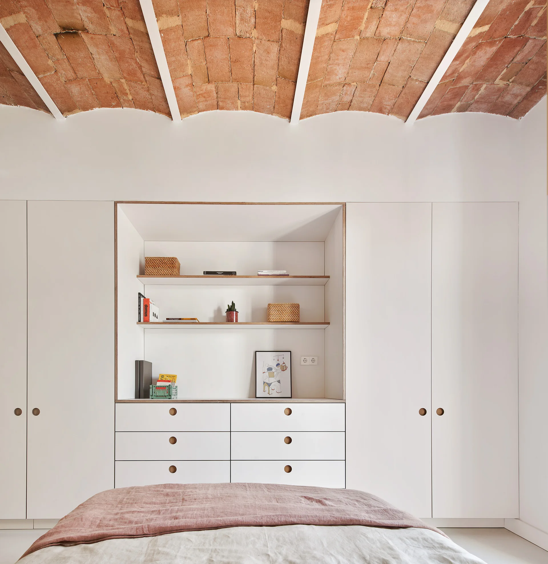 A bedroom features a minimalist white storage wall with built-in shelves and drawers beneath an arched terracotta brick vaulted ceiling.