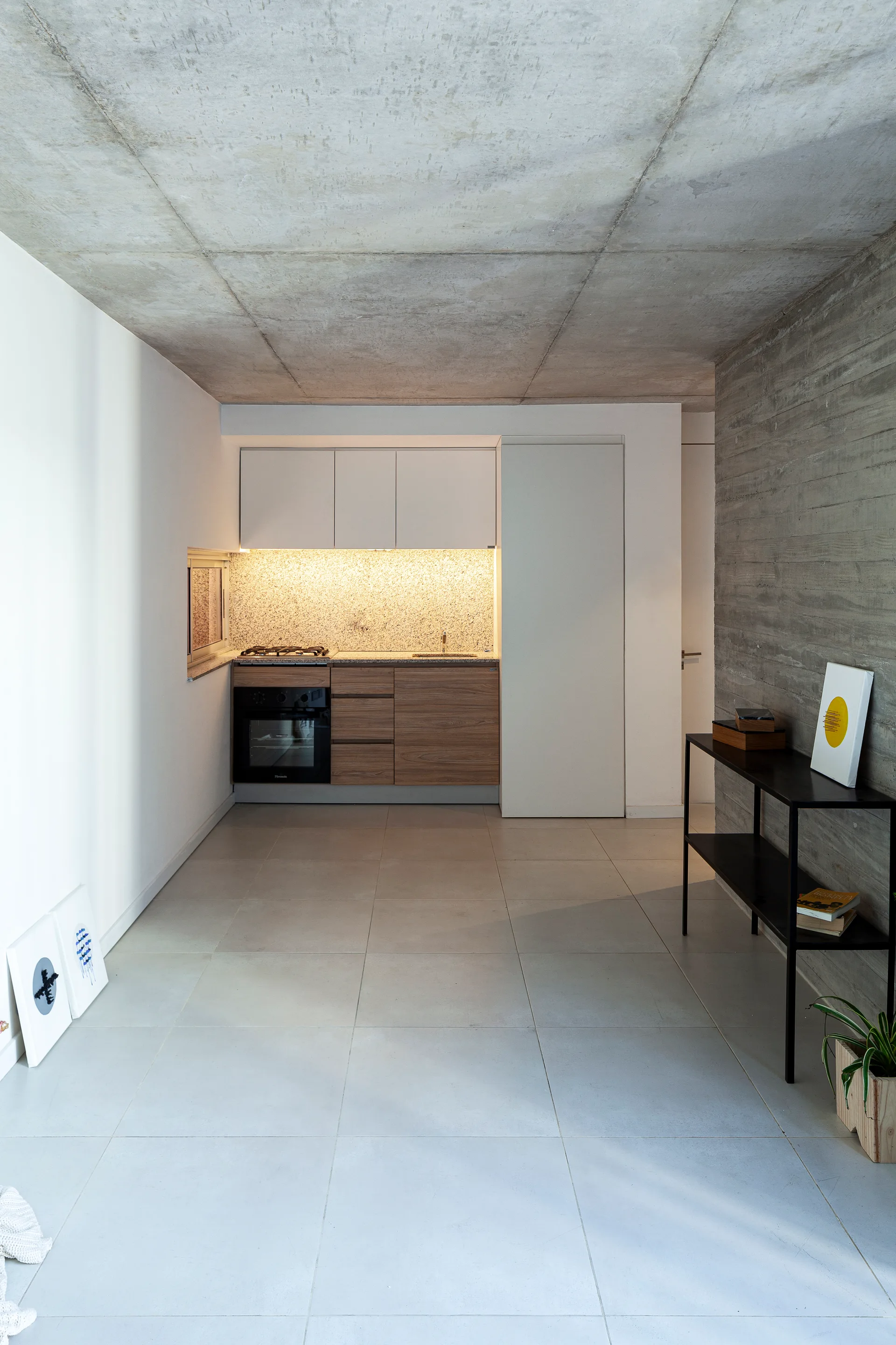 A minimalist kitchen features a raw concrete ceiling, wooden lower cabinets, white upper cabinetry, and a black console table against a textured concrete wall.