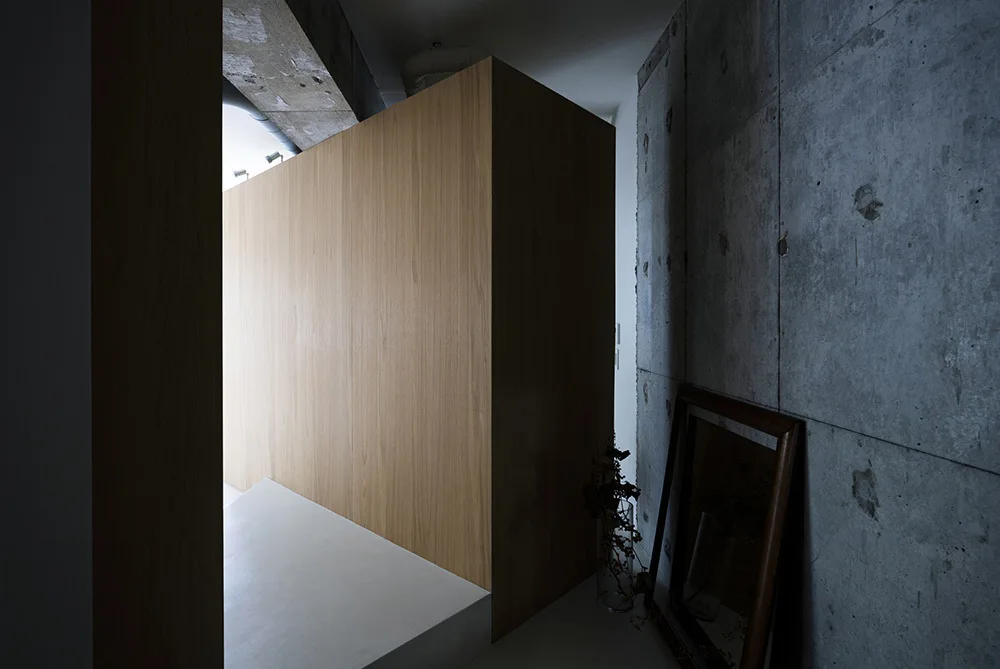 A large light wood partition wall stands next to an exposed concrete wall with a leaning wood framed mirror and a glass vase.