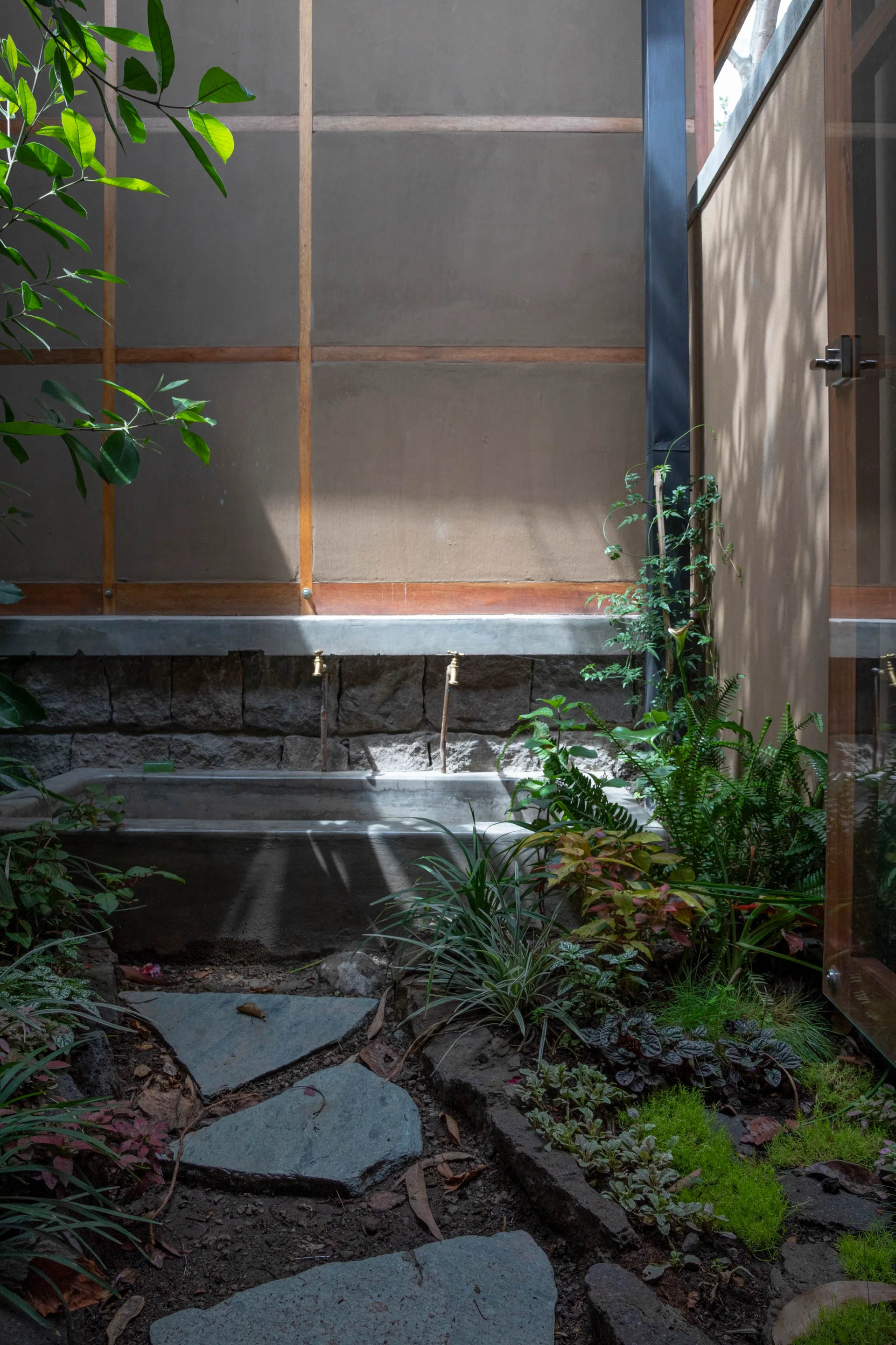 An outdoor courtyard with a concrete wash basin, brass faucets, rough stone wall, slate stepping stones, and lush green plants.