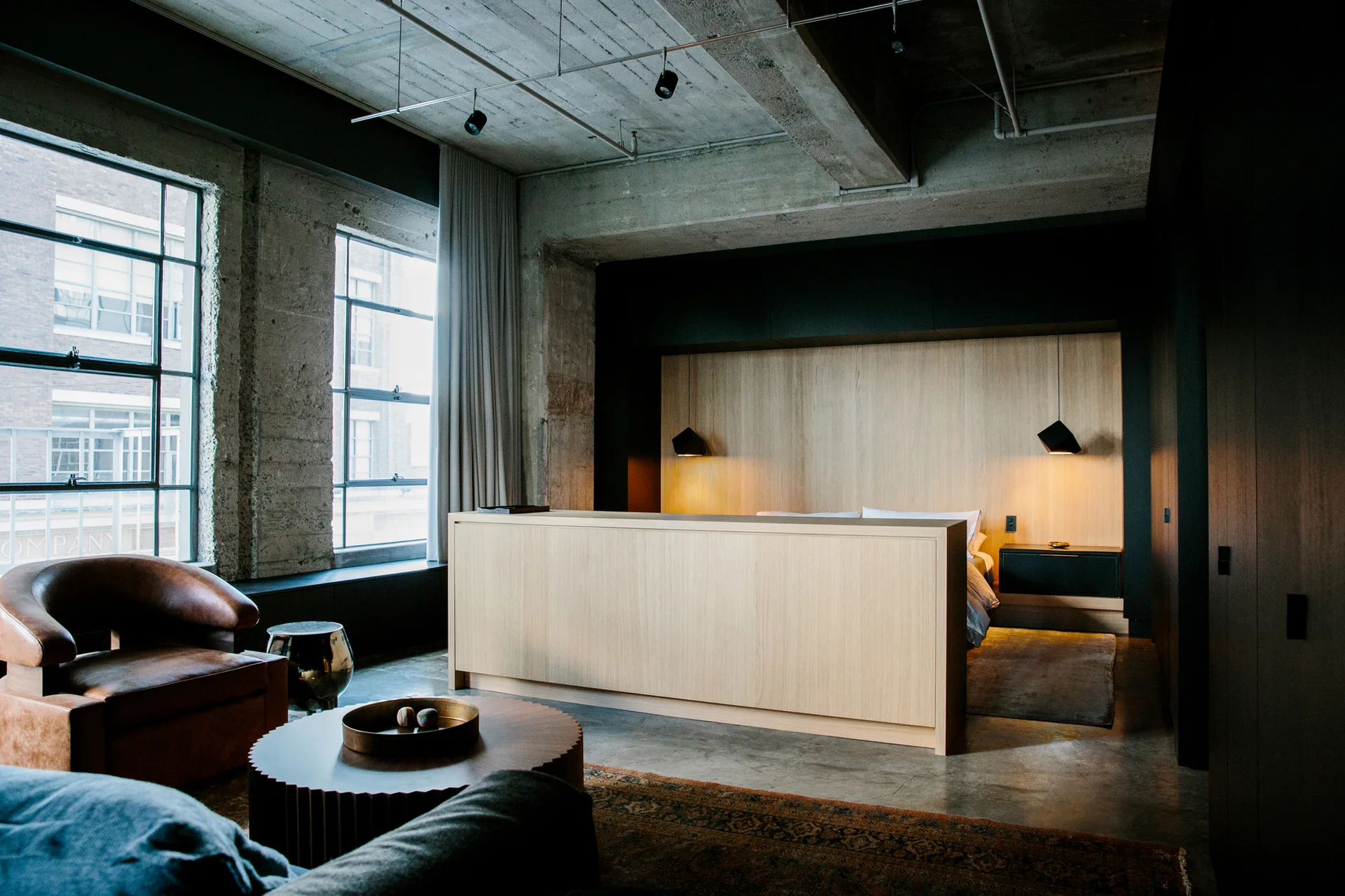 An industrial loft bedroom features an exposed concrete ceiling, a light wood partition divider, a leather armchair, and large grid windows.