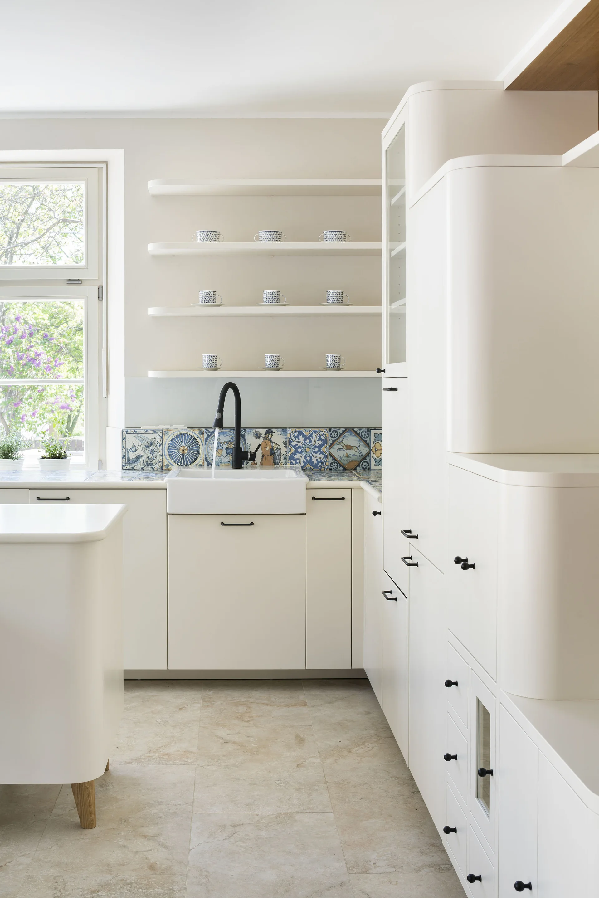 A bright white kitchen featuring cabinetry with curved edges, a farmhouse sink with a black faucet, and a decorative blue patterned tile backsplash.