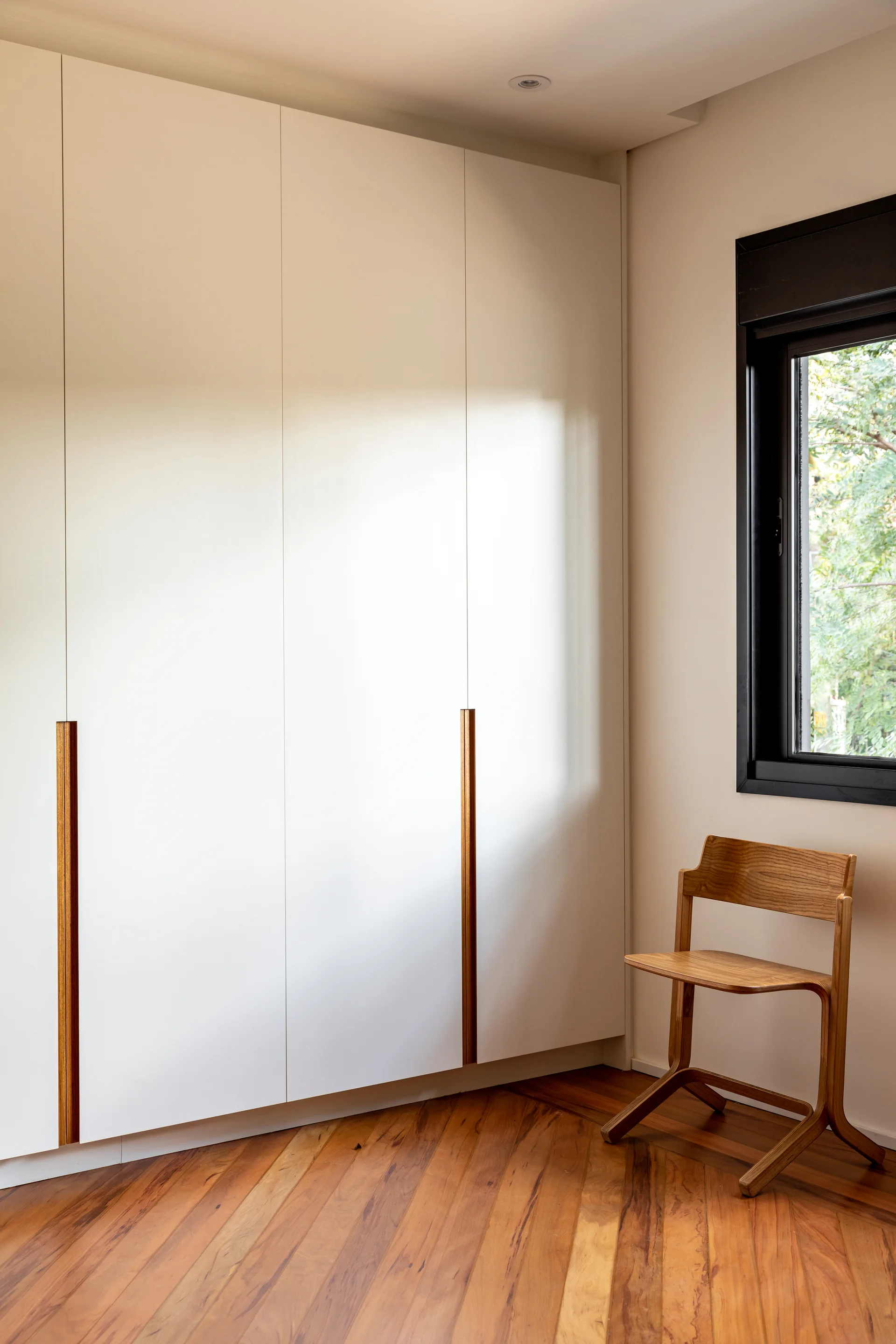 A minimalist white built-in wardrobe with vertical wood pulls stands next to a wooden chair on diagonal hardwood floors in a sunlit corner.