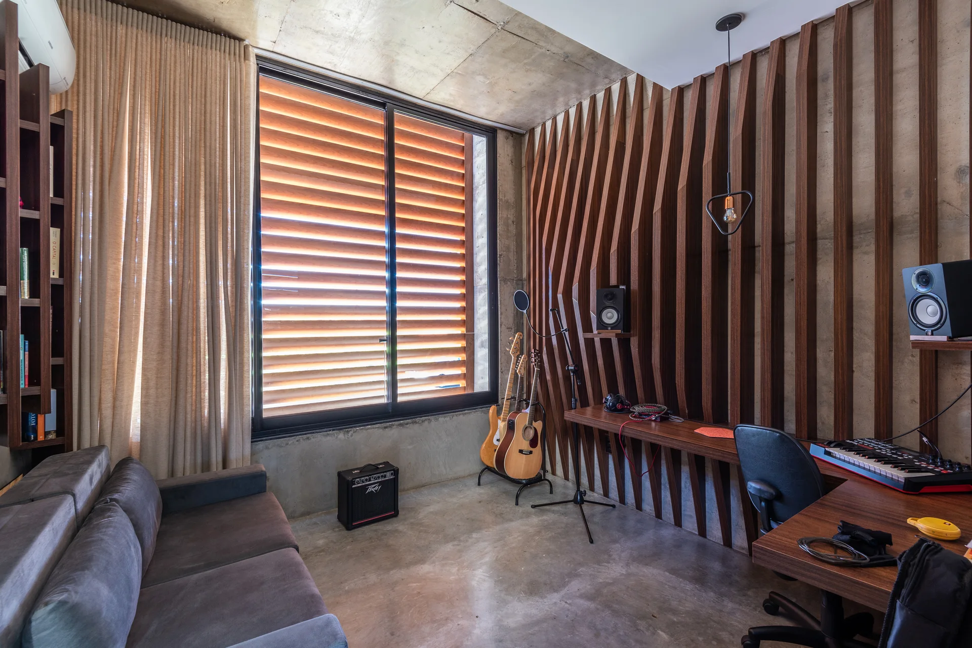 A home music studio features a vertical wood slat accent wall, a built-in wooden desk, concrete walls, and a large window with louvers.