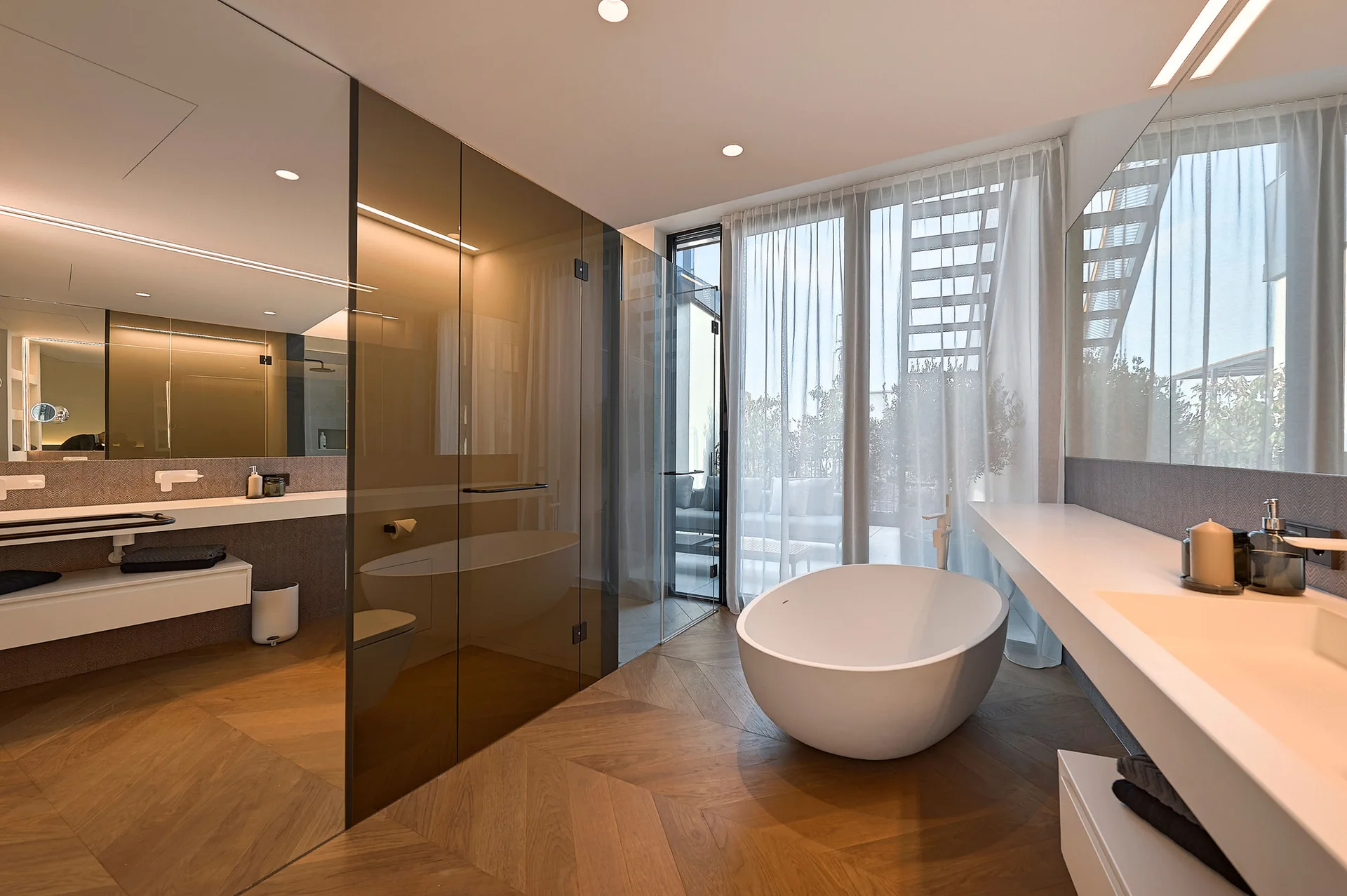 A modern bathroom with chevron pattern wood flooring, a white freestanding tub, a long floating vanity, and tinted glass shower partitions.