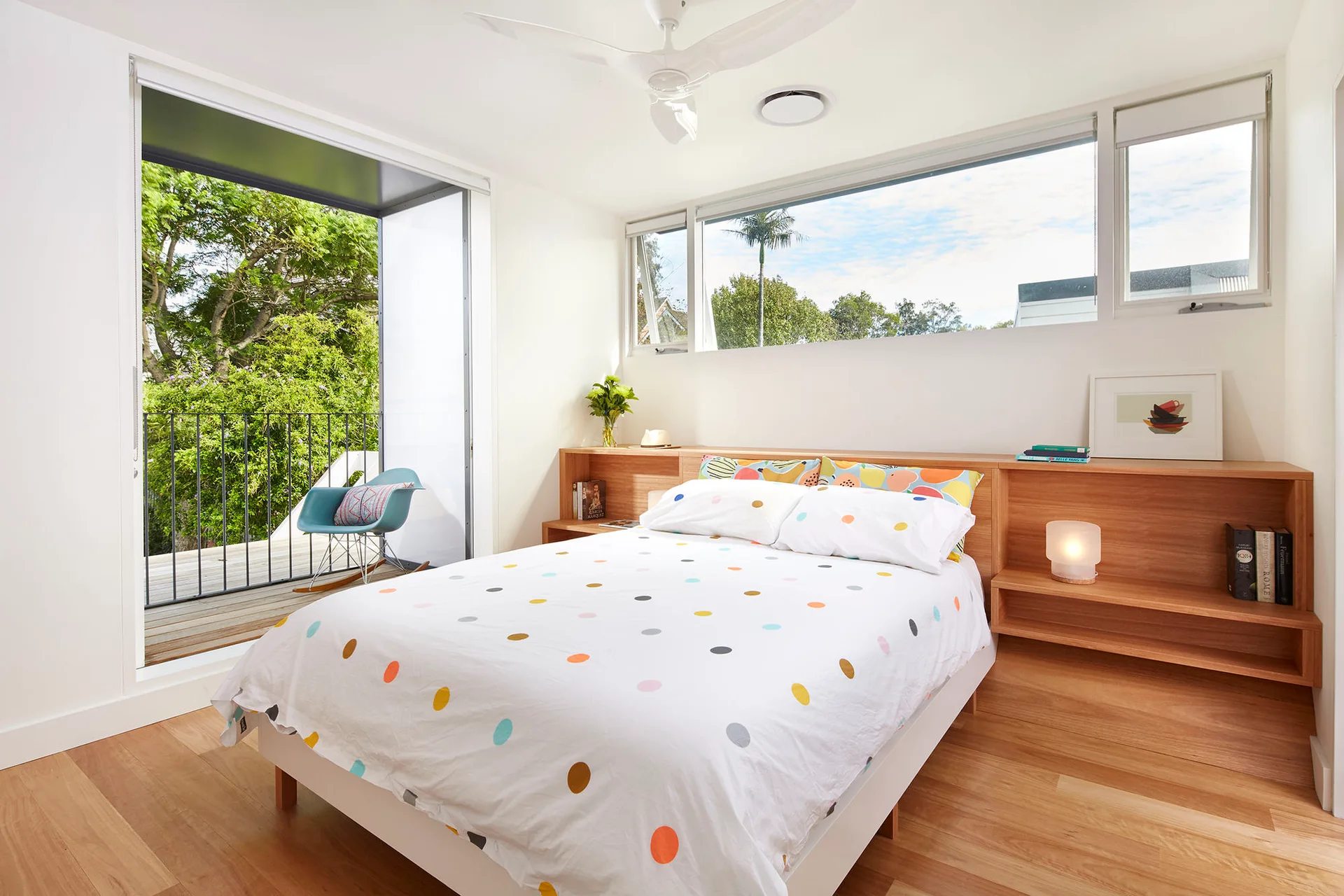 A modern bedroom features a low bed with polka-dot bedding set against a custom built-in wooden headboard with integrated shelves.