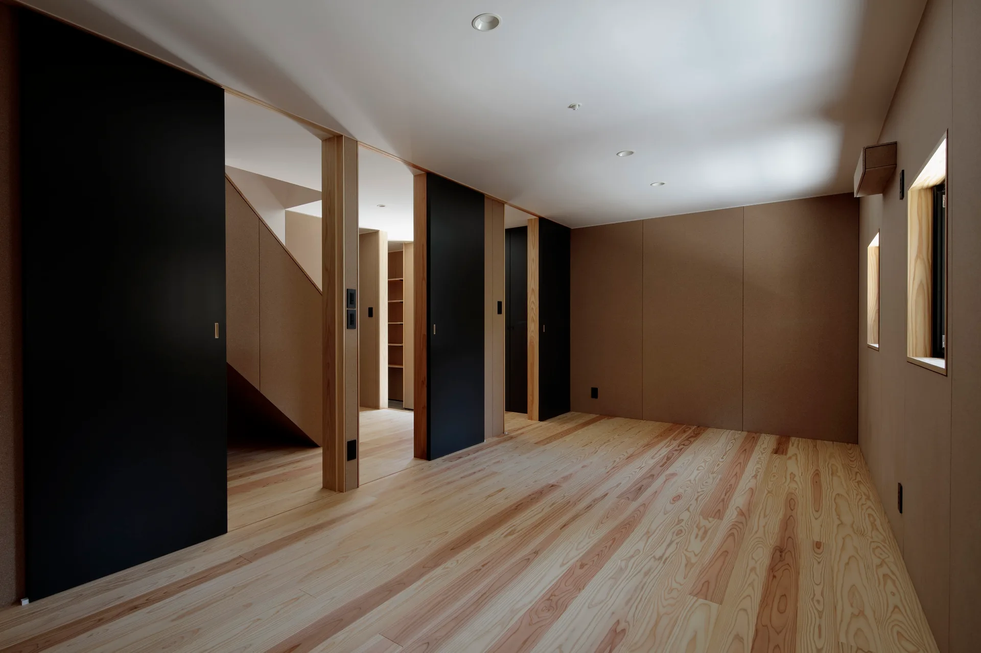 A minimalist hallway features light wood plank flooring, textured brown wall panels, and matte black sliding doors beneath a white ceiling with recessed lights.