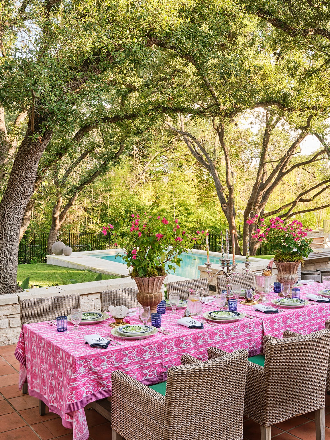 An outdoor dining table with a pink ikat tablecloth and wicker chairs sits on a terracotta patio under large shade trees near a pool.