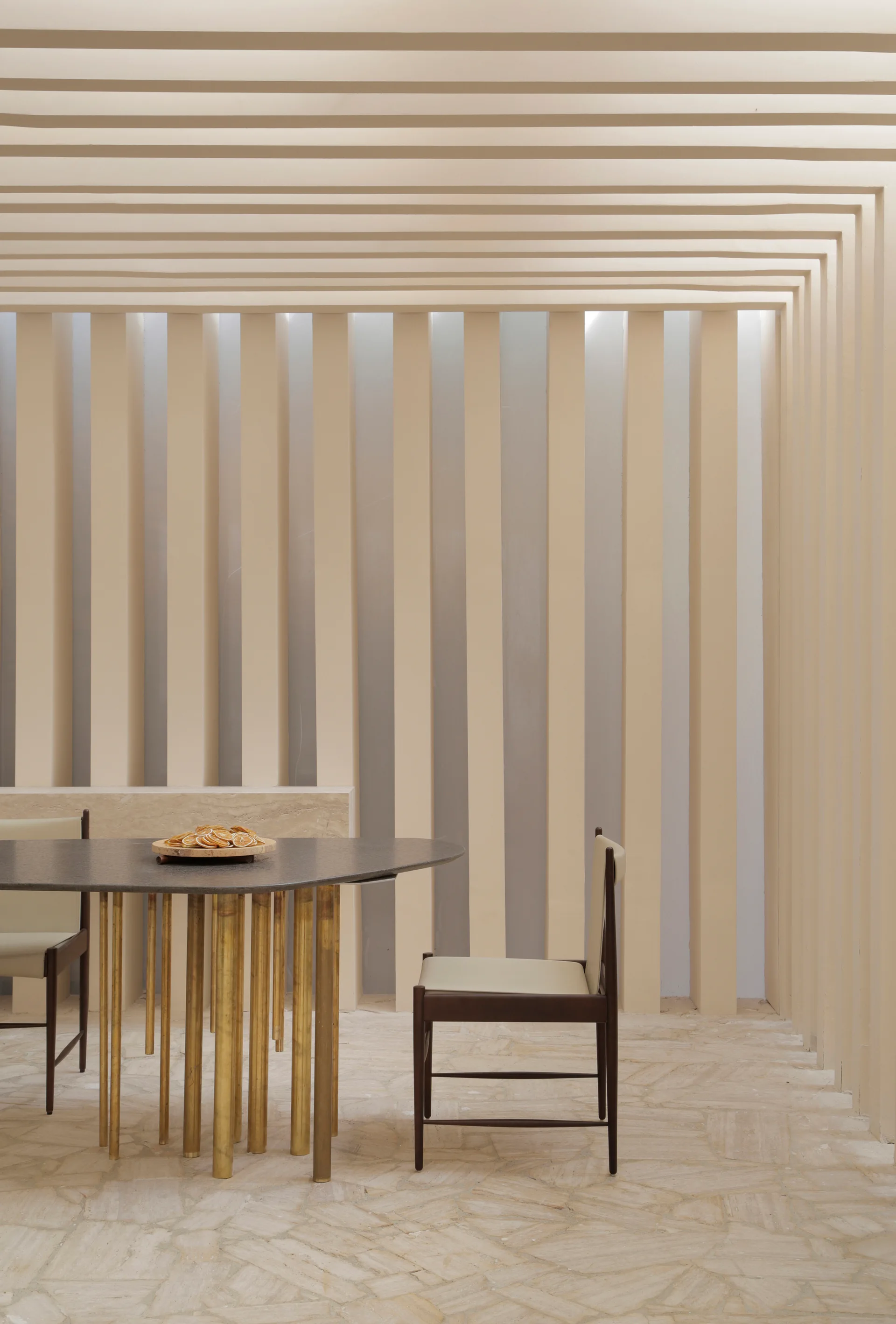A dark-topped dining table with brass cylindrical legs is paired with wood chairs in front of a light cream slatted wall and flagstone floor.