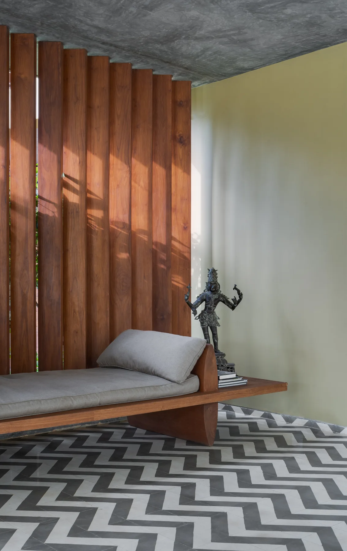 A low wooden daybed with a gray cushion sits against a vertical wood slat partition on a black and white chevron tiled floor.