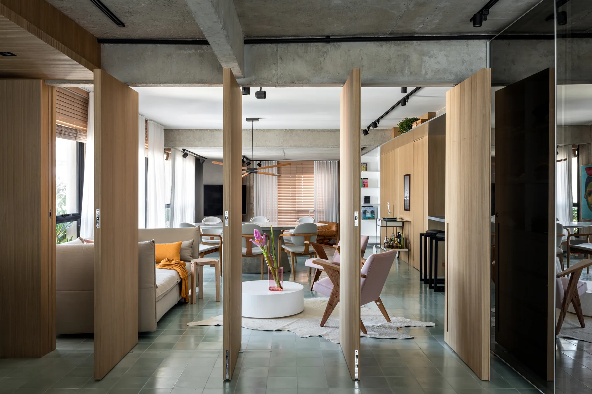 A contemporary living room features warm wood pivoting partition doors, a raw concrete ceiling, green floor tiles, and mid-century furniture.