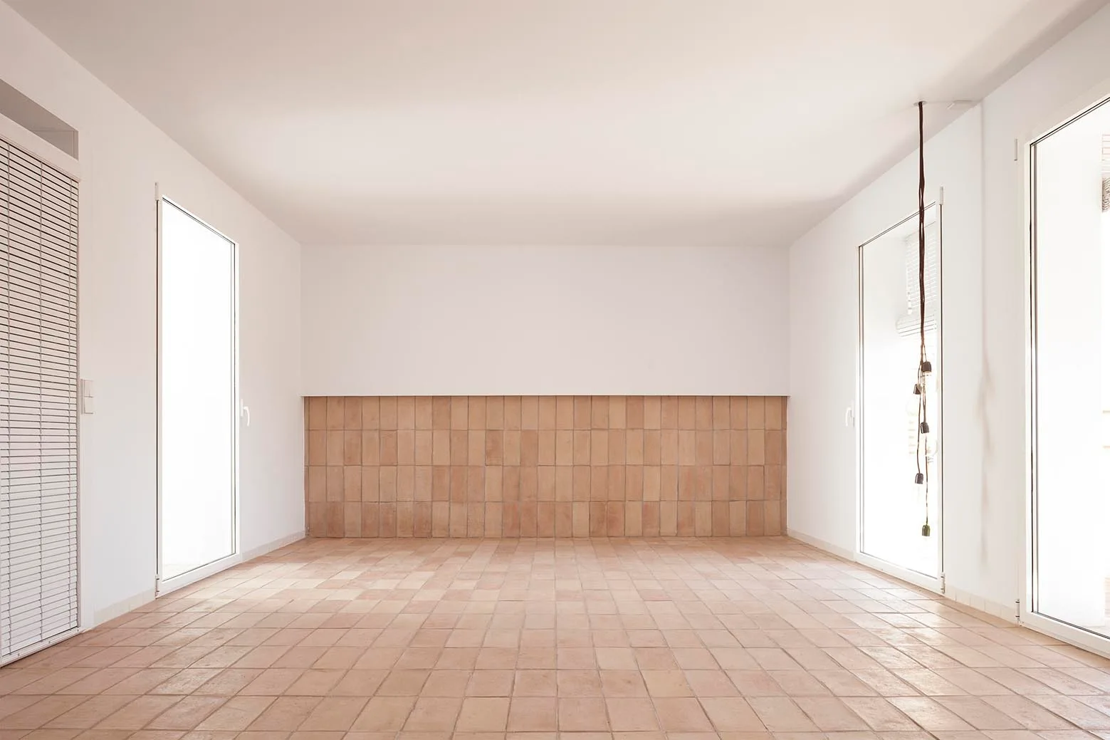 An empty minimalist room featuring warm terracotta floor tiles, a matching tile wainscot on the back wall, and bright windows on both sides.