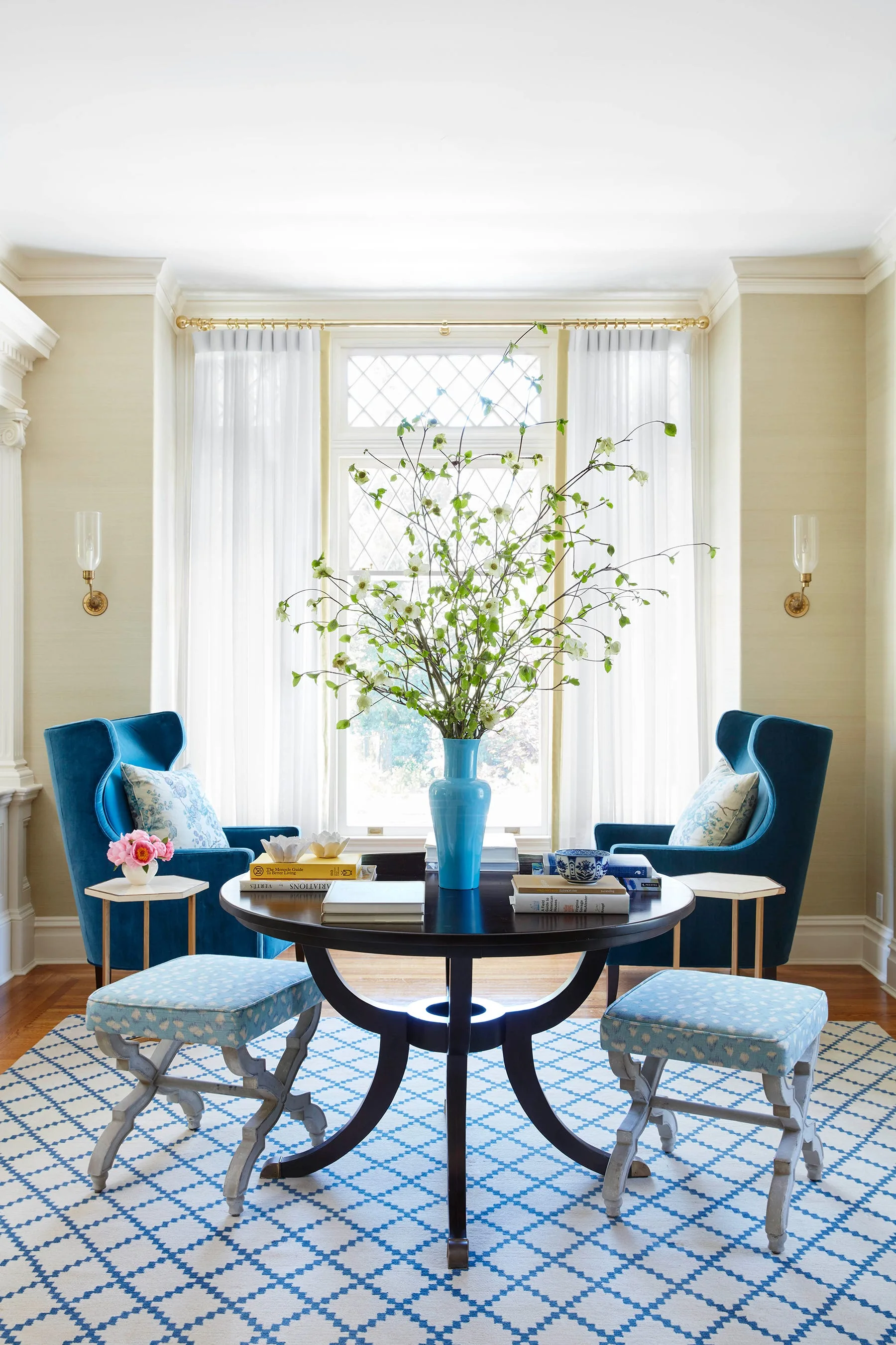 Two blue velvet wingback chairs flank a round dark wood table with a tall branch centerpiece in front of a bright window.