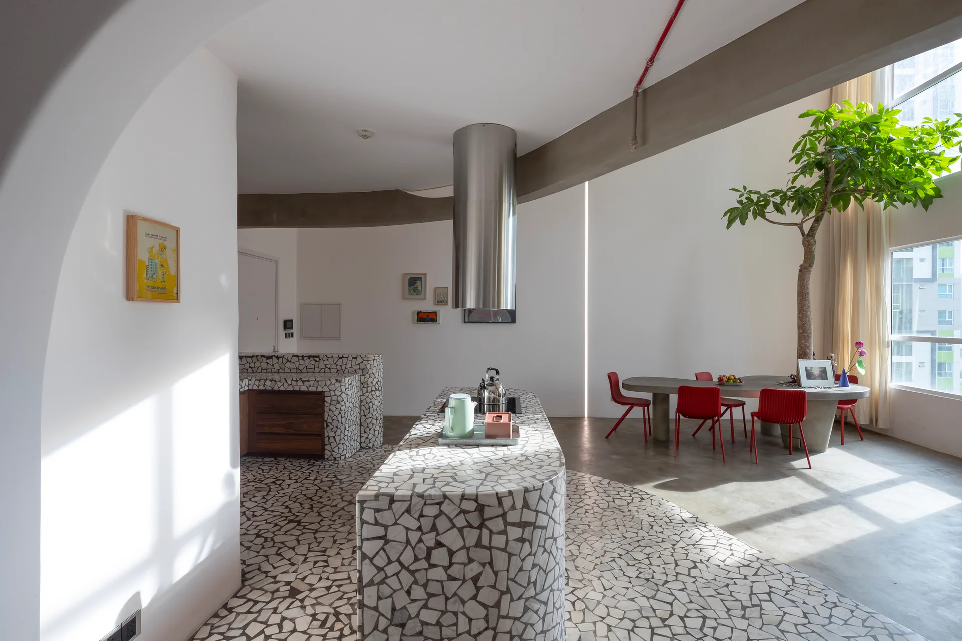 An open-concept kitchen features a curved mosaic-tiled island, a cylindrical range hood, and a dining table surrounded by red chairs.