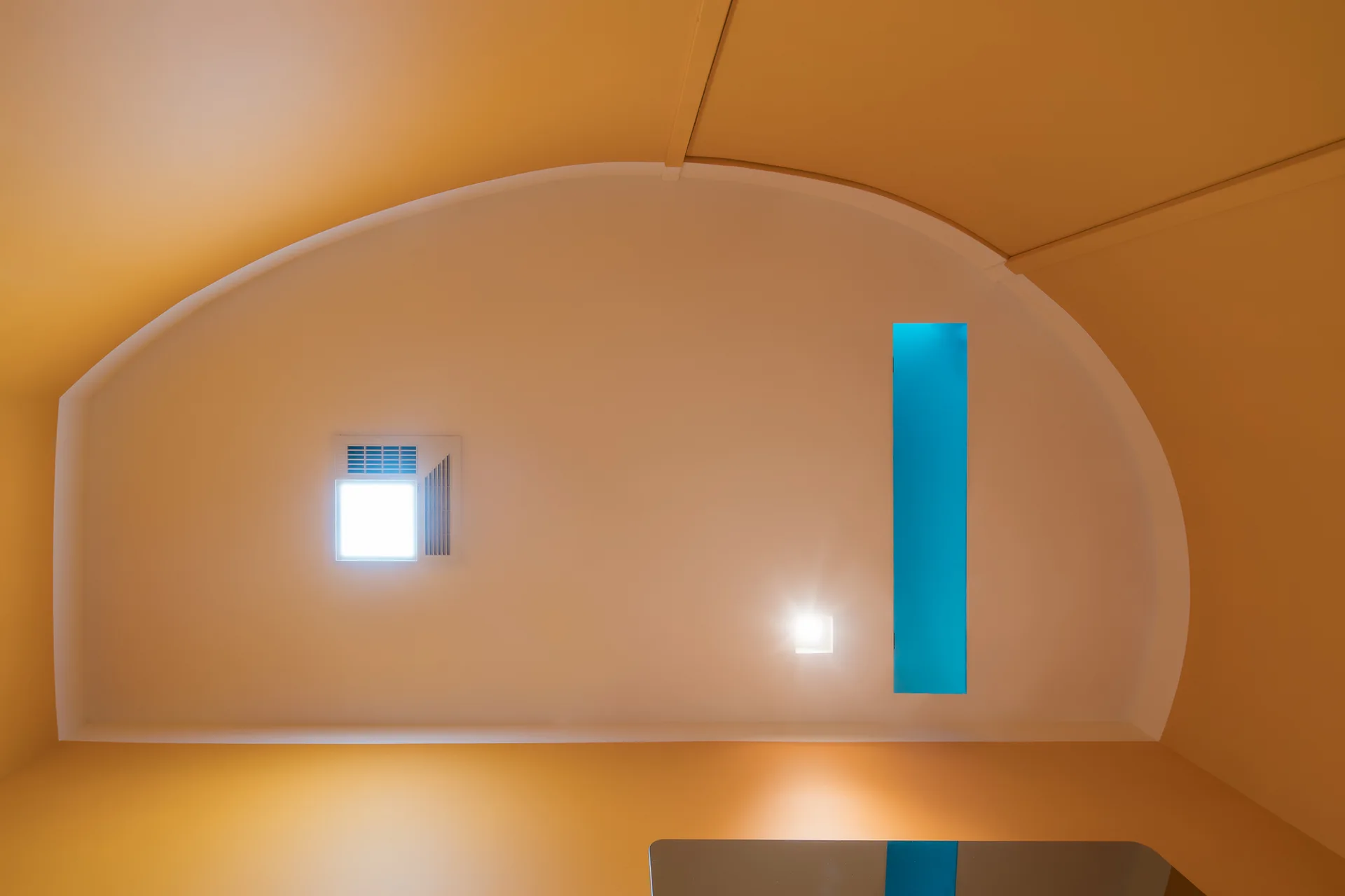 A low-angle view of a curved white ceiling framed by yellow walls, featuring two recessed square lights and a rectangular blue insert.