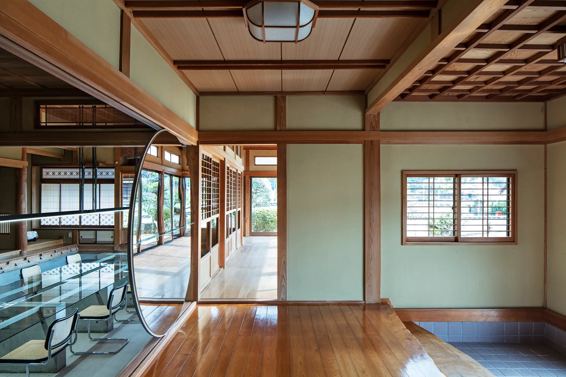 A traditional Japanese room features natural wood framing, a coffered ceiling, and a modern glass dining table behind a circular metal archway.