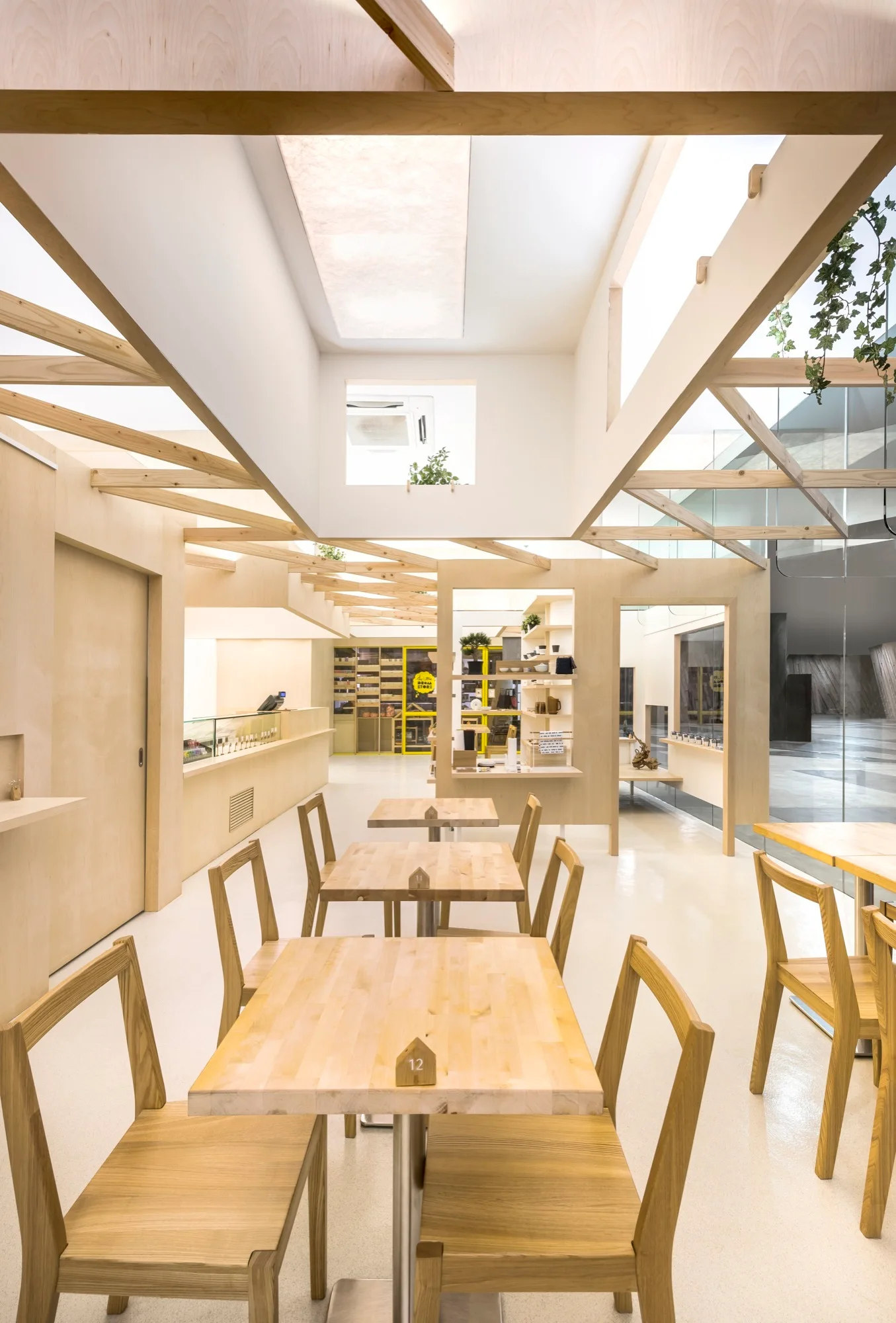 A bright minimalist cafe featuring rows of light wood tables and chairs under a ceiling with exposed timber beams and skylights.