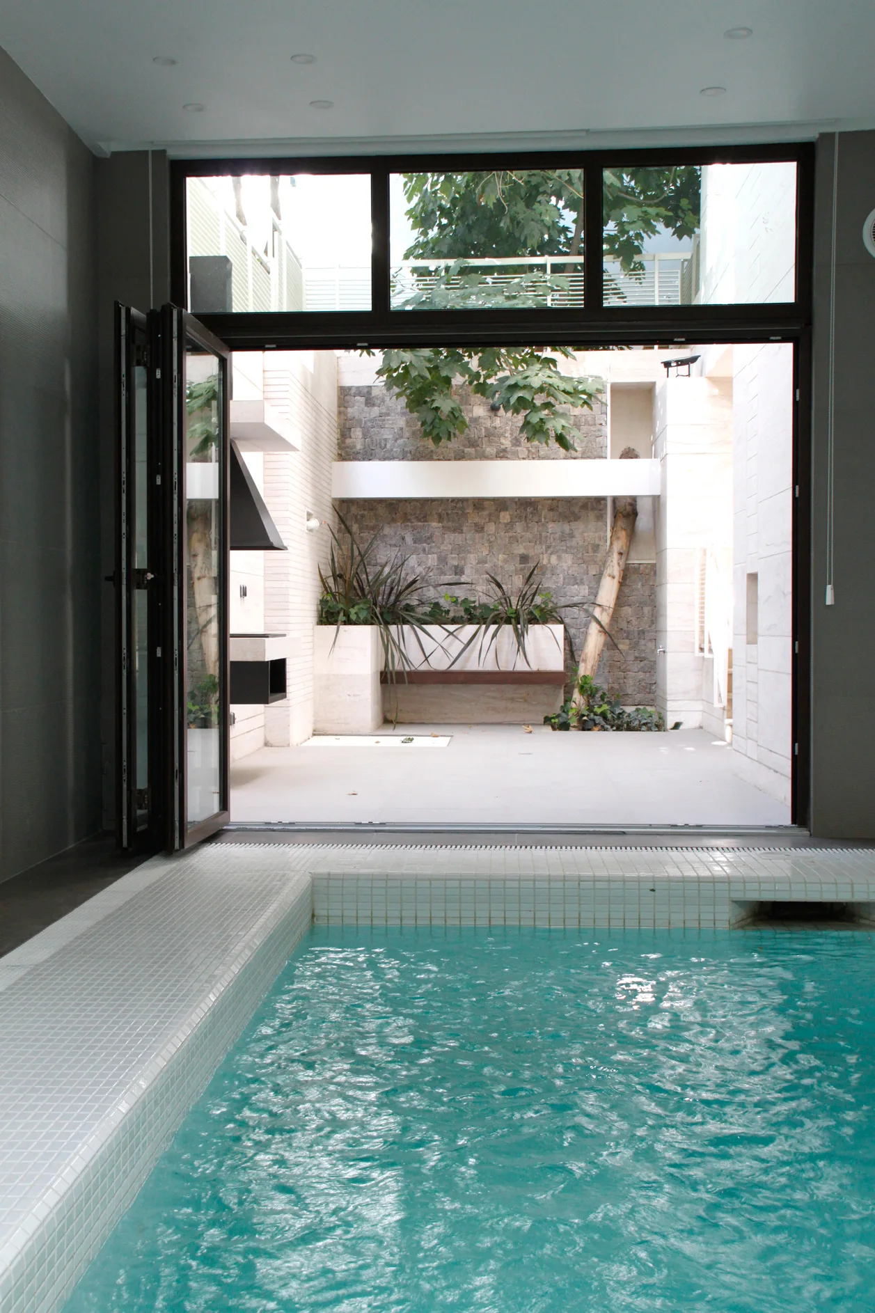 An indoor swimming pool with turquoise water and white mosaic tile borders sits next to open black-framed folding glass doors leading to a stone patio.