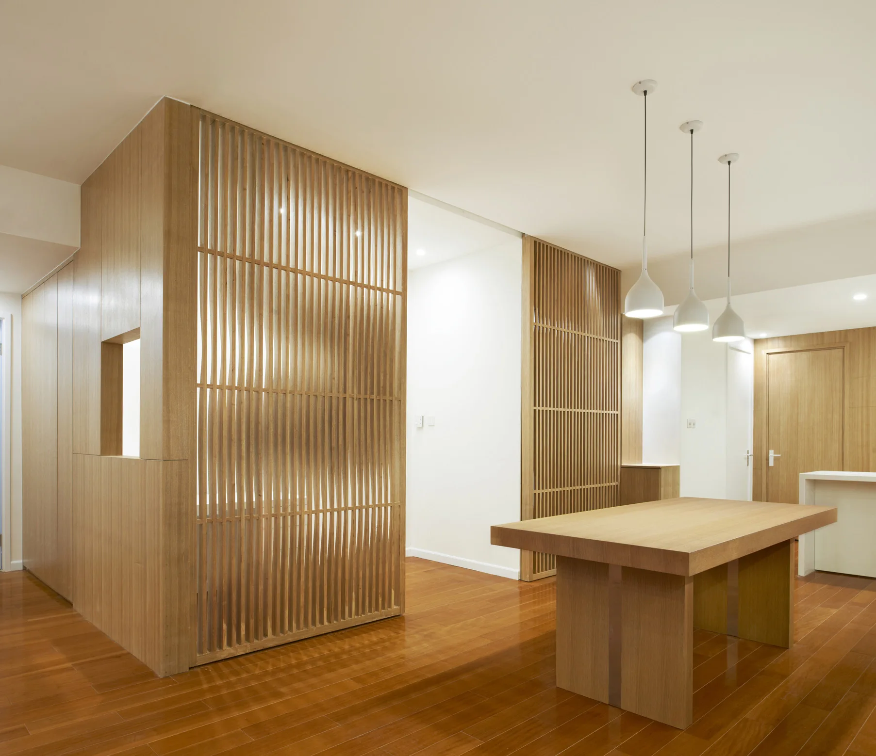 A minimalist dining area featuring a long wood table, three white pendant lights, and floor-to-ceiling vertical wood slat partition screens.