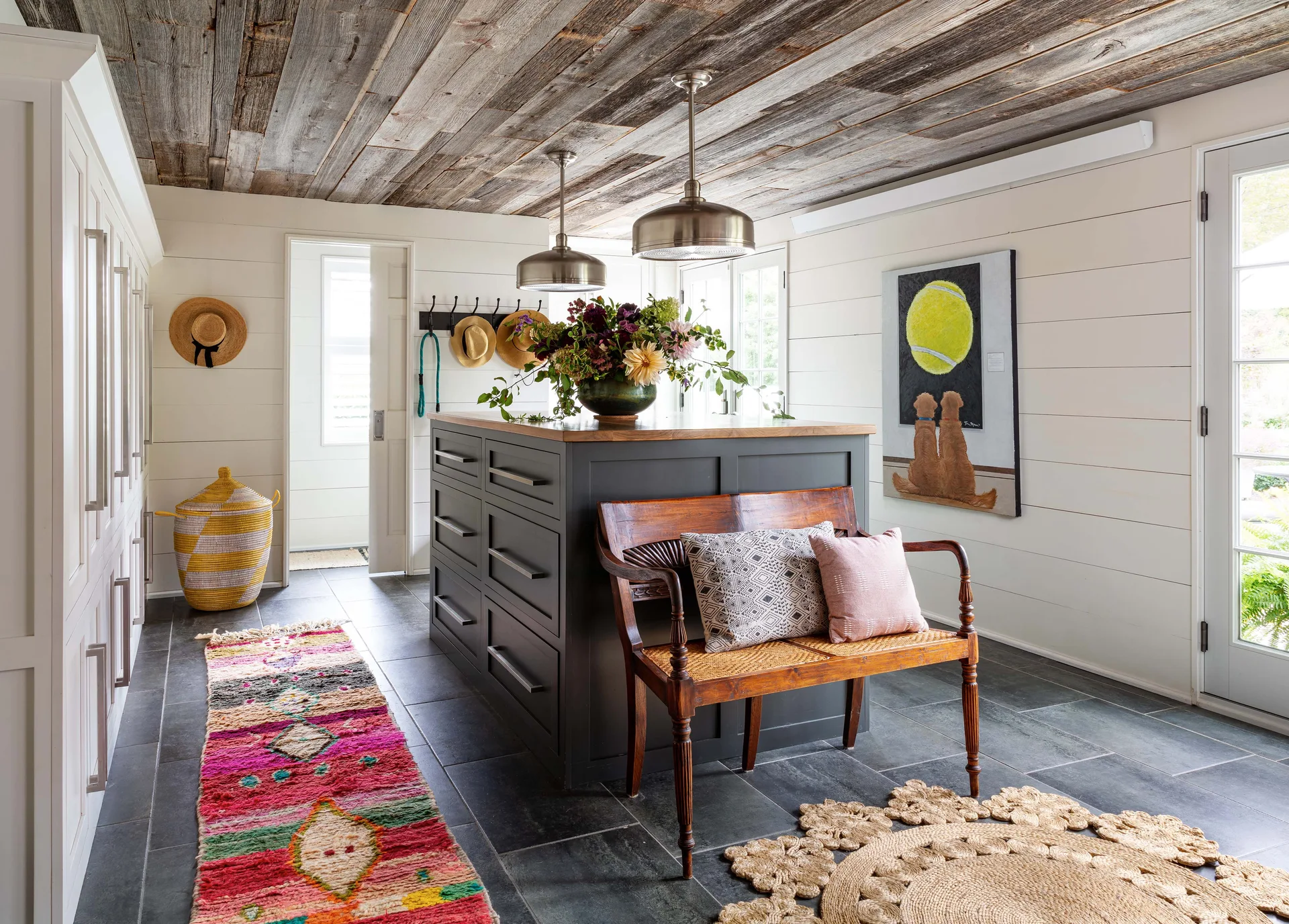 A farmhouse mudroom featuring a dark gray island, a vintage wooden bench, white shiplap walls, and a reclaimed wood plank ceiling.