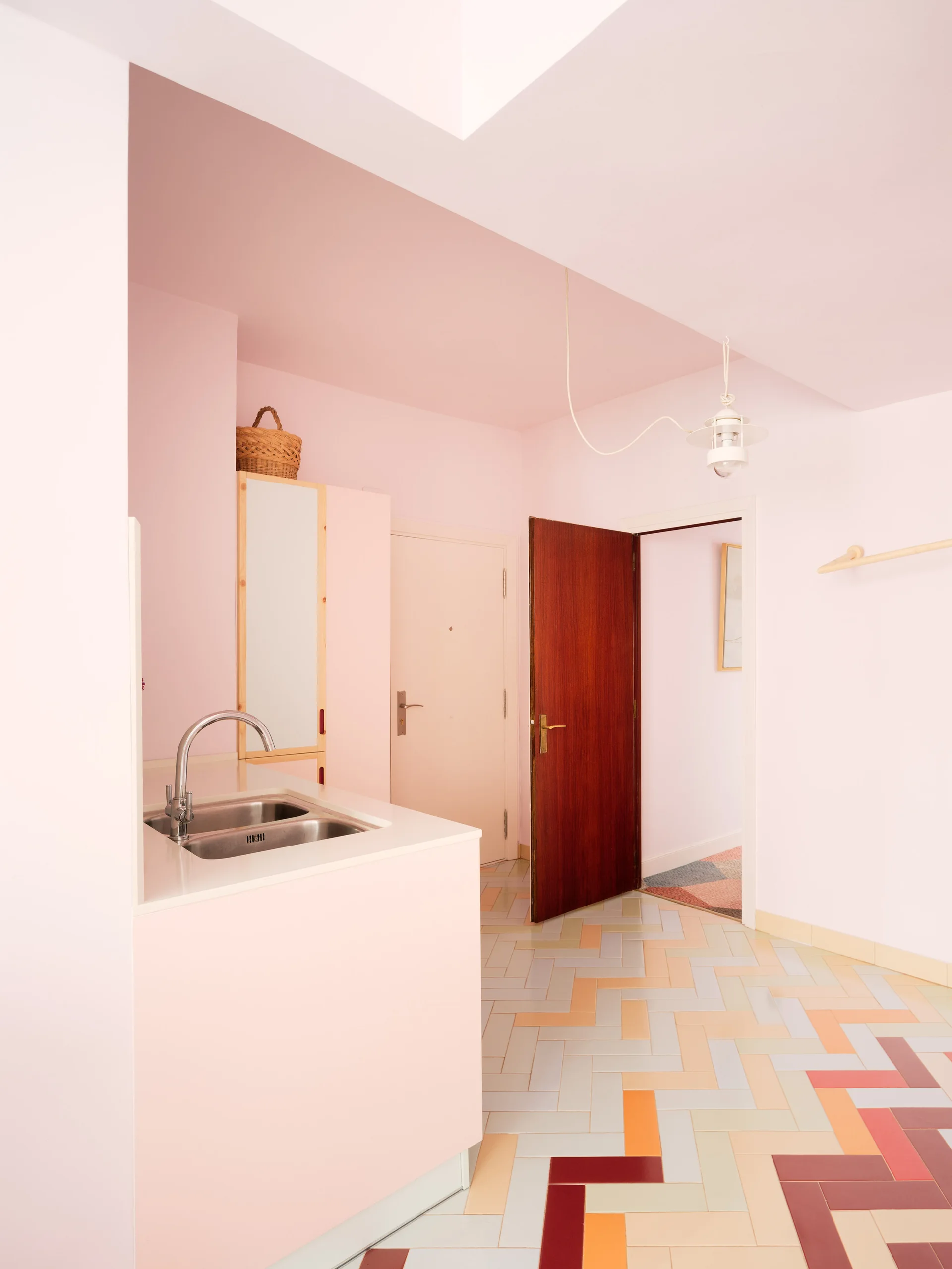 A minimalist kitchen featuring soft pink walls and cabinetry paired with a sink and multicolored herringbone floor tiles.