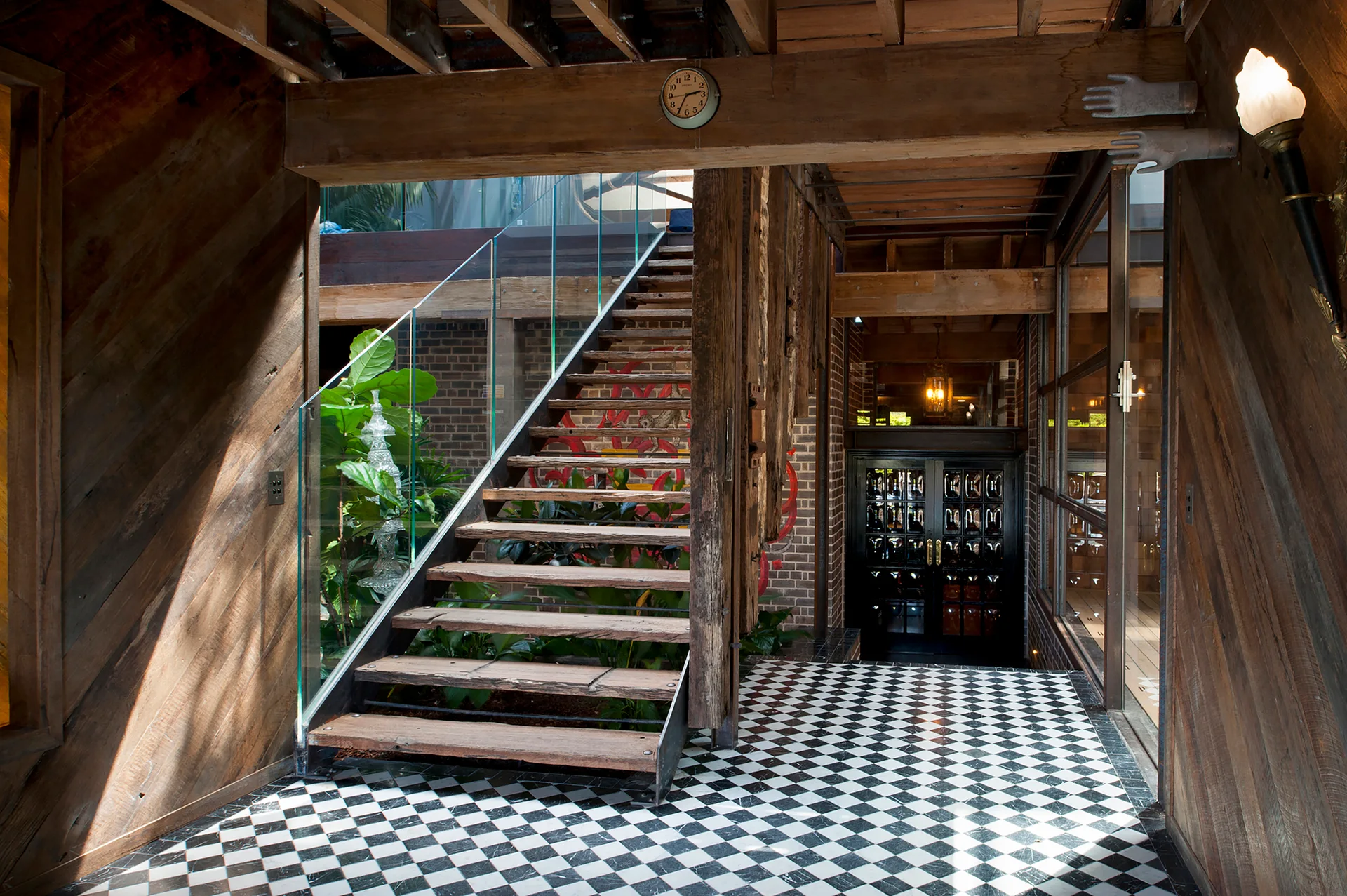A rustic entryway features an open-tread wood staircase with a glass railing, set on a black and white checkerboard tile floor under exposed ceiling beams.