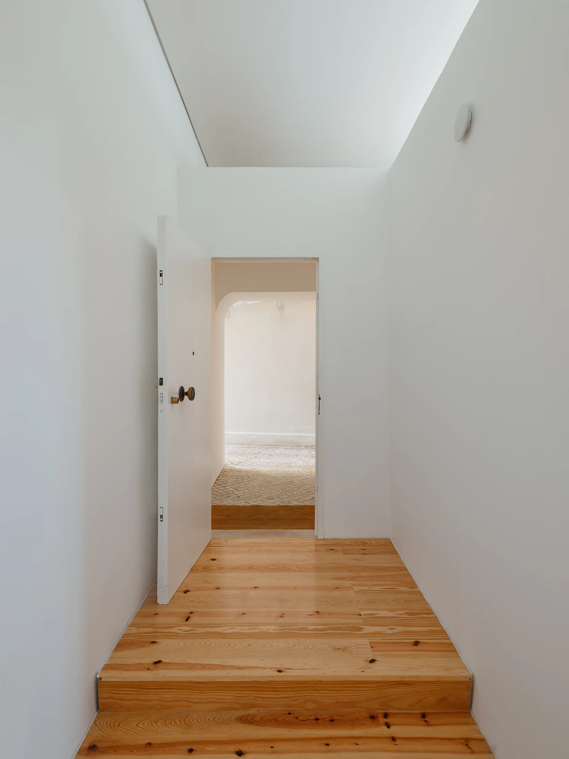A minimalist white entryway features wide knotty pine floorboards, a short step, and an open white door leading to a sunny outdoor area.