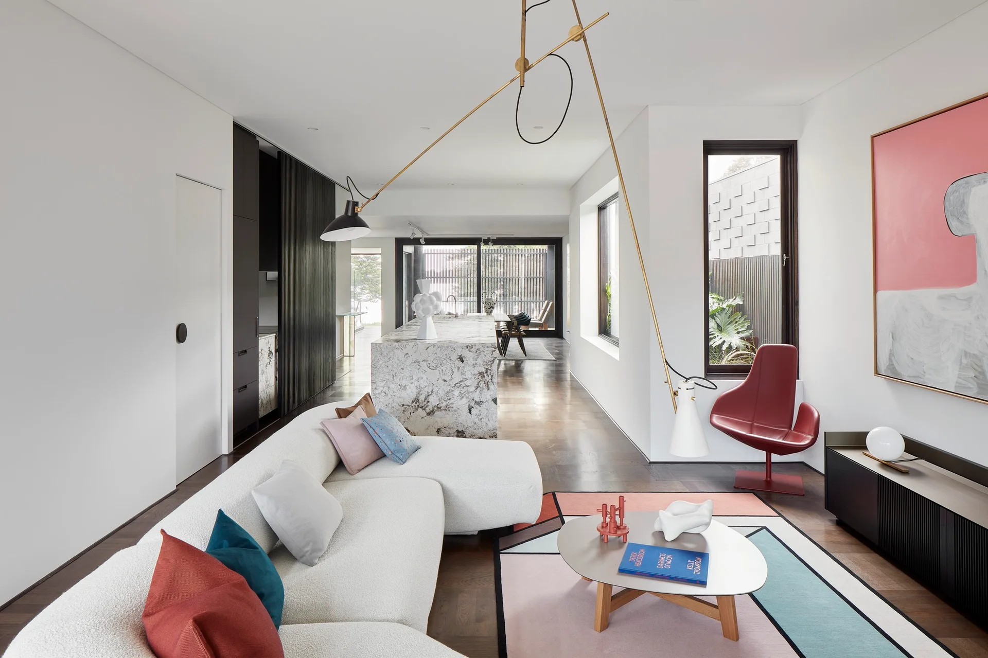 A bright open-plan living room featuring a curved white sofa on a colorful geometric rug, with a large stone island and swing-arm floor lamp.
