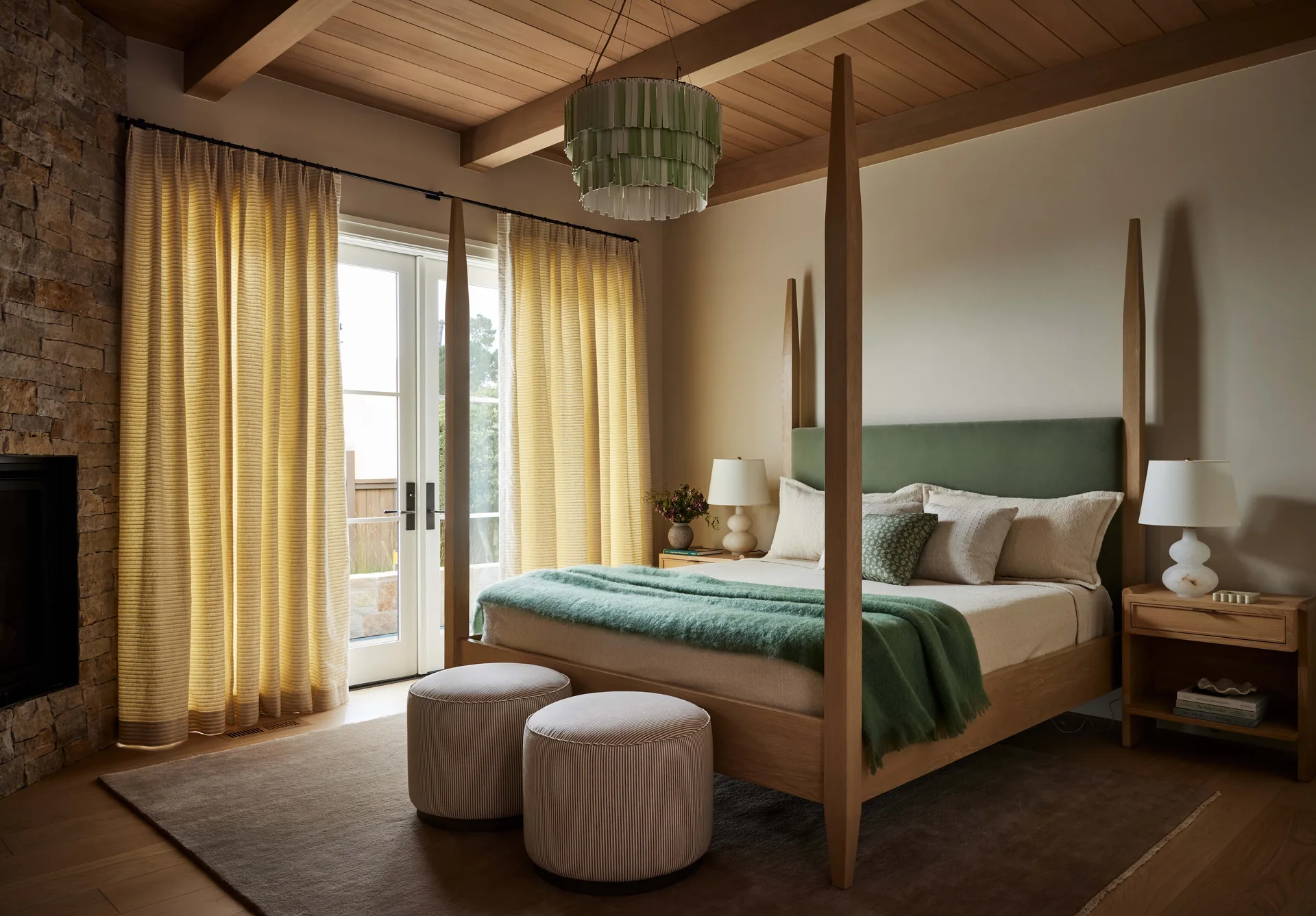 A wooden four-poster bed with a green headboard stands in a bedroom featuring wood-beamed ceilings, linen drapes, and a stone fireplace.