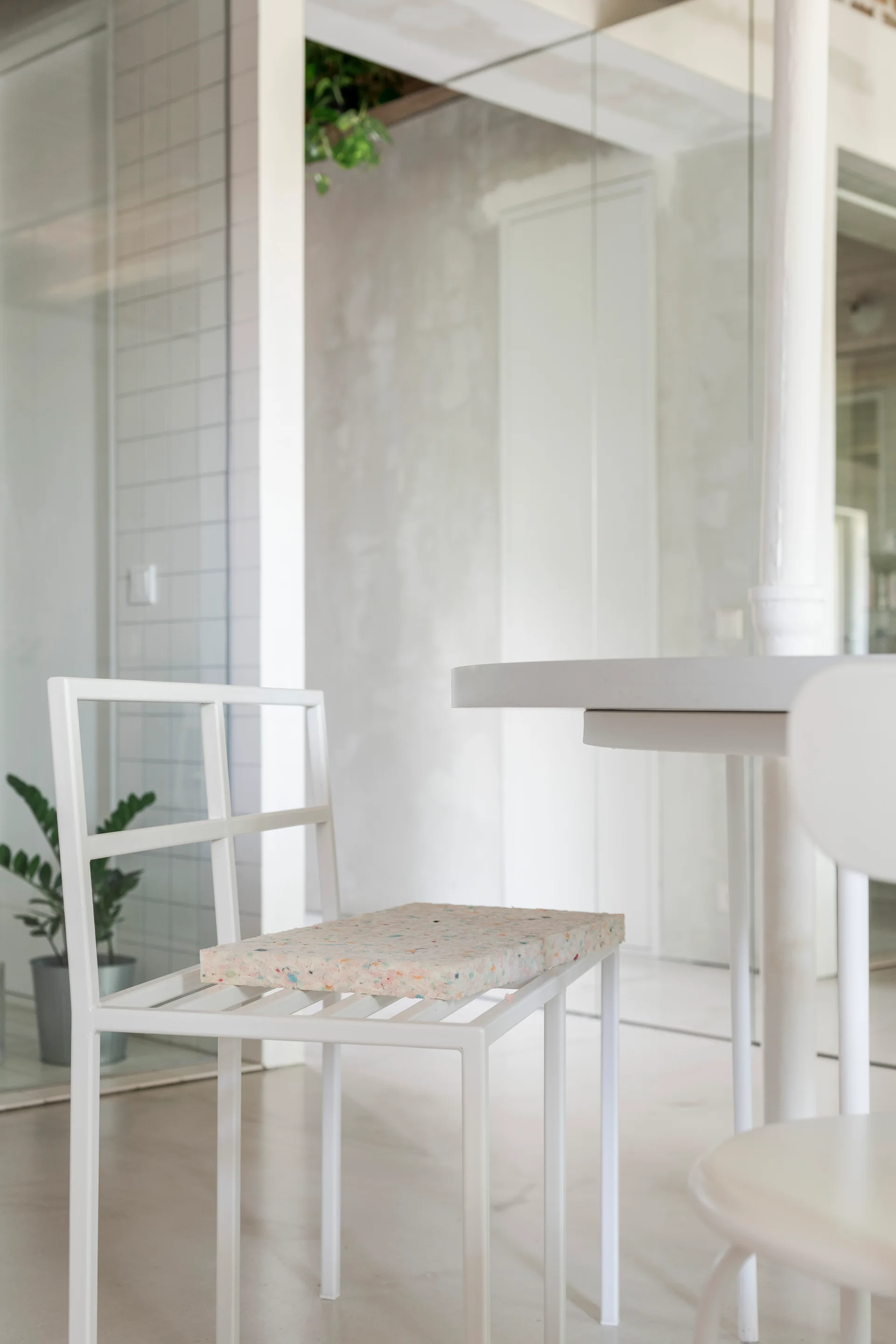 A white geometric metal dining chair features a multicolored speckled foam cushion next to a round white table in a minimalist grey concrete room.