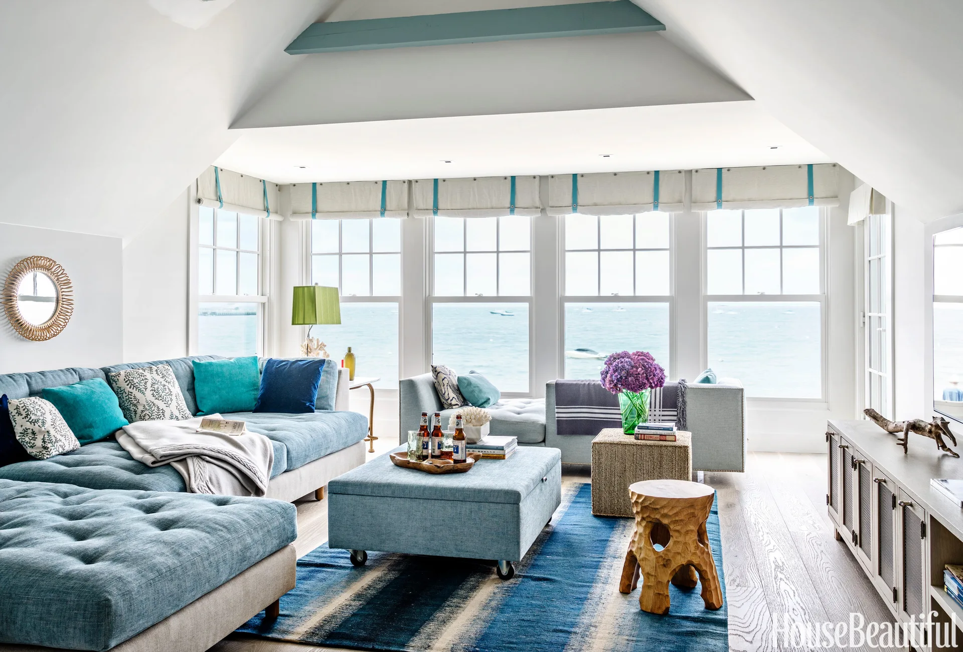 A bright coastal living room features a blue sectional sofa, a matching fabric ottoman, and large windows looking out over the ocean.