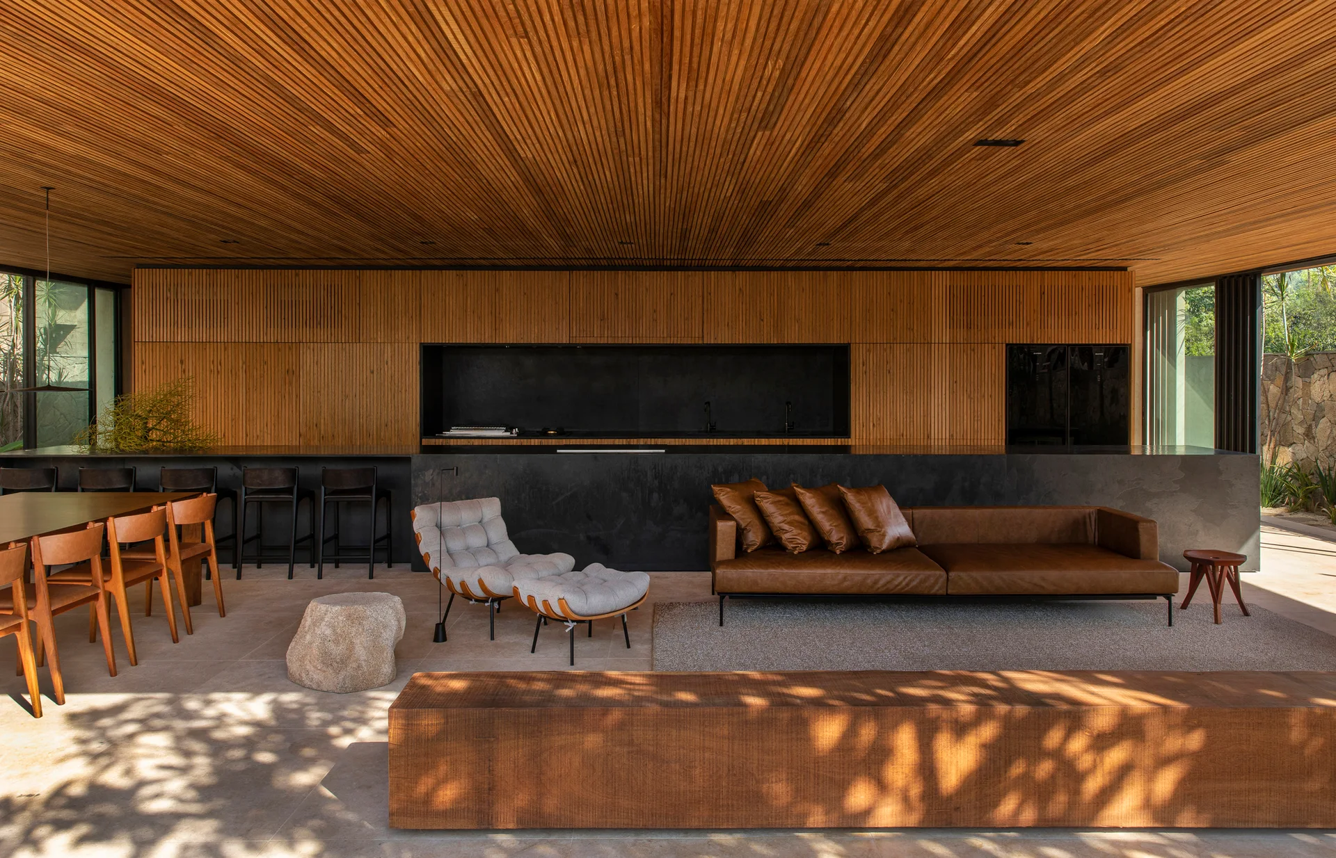 A modern open-plan living room and kitchen featuring slatted wood ceilings and walls, a black stone kitchen island, and a brown leather sofa.