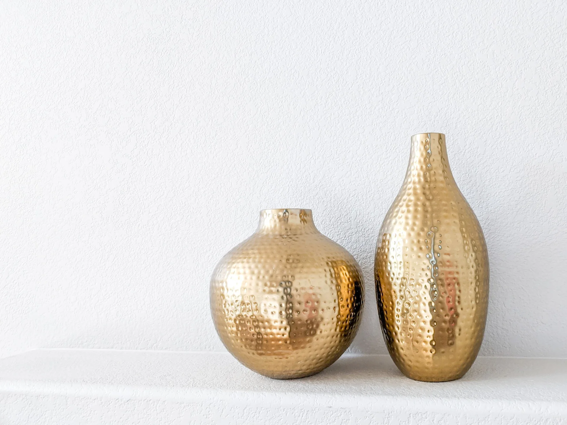 Two gold hammered metal vases of varying heights rest on a white plaster ledge against a textured white wall.