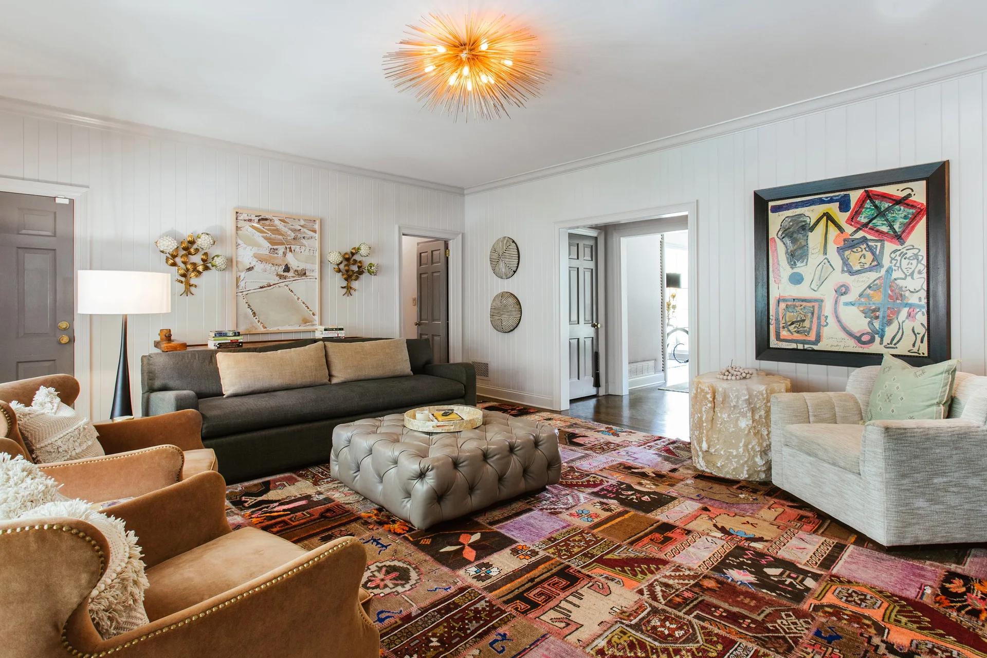 A spacious living room features a colorful patchwork rug, a grey tufted ottoman, velvet armchairs, and vertical white wall paneling.
