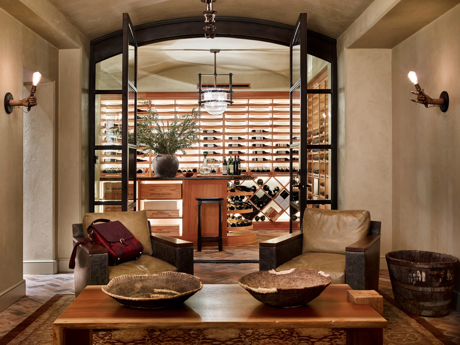 A wine cellar with backlit wooden racks is viewed through arched glass French doors from a lounge featuring leather armchairs and a brick floor.