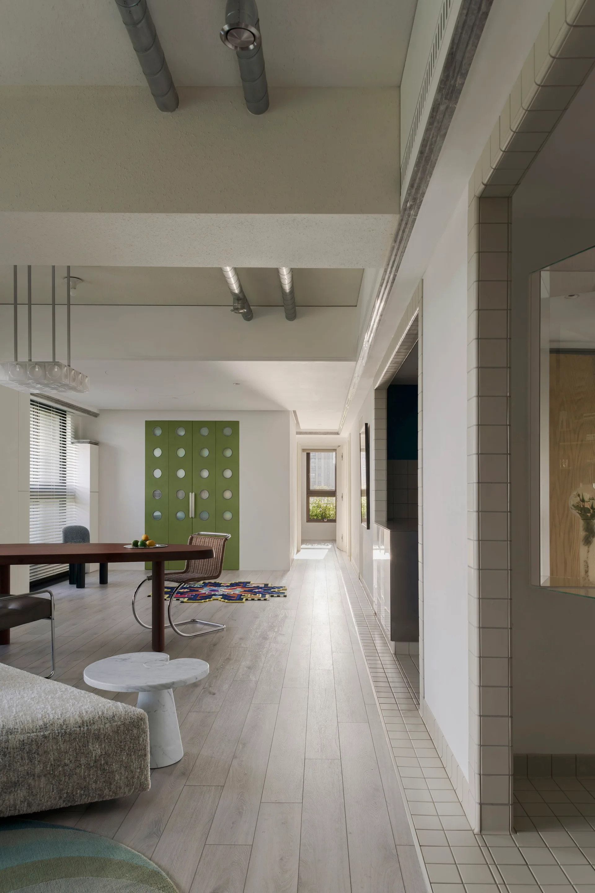 A light-filled dining space features a wooden table, a green double door with circular glass inserts, and exposed silver ceiling ducts.