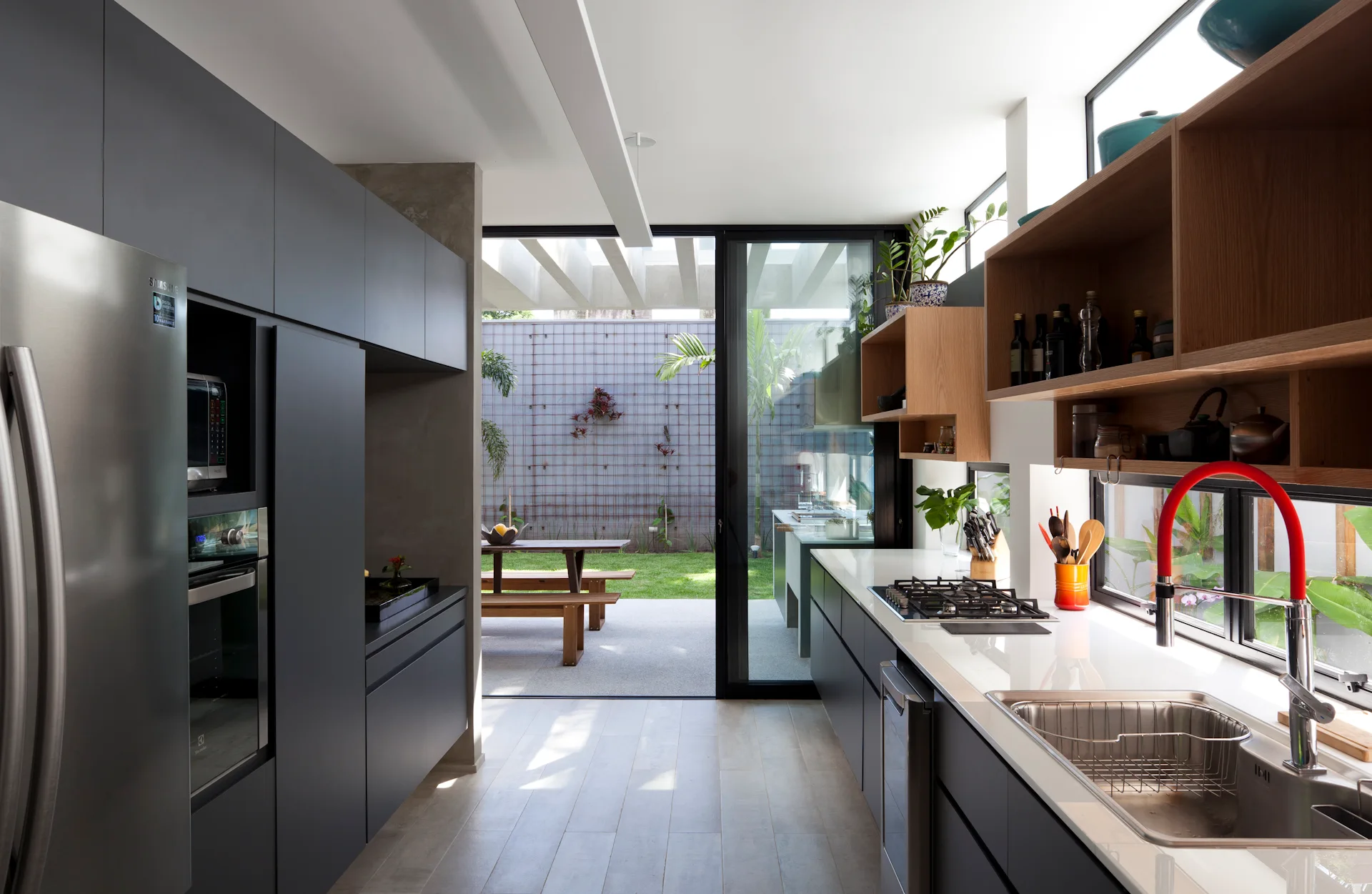 A modern galley kitchen features matte grey cabinets, floating wooden shelves, a red faucet, and a glass door opening to a green backyard.