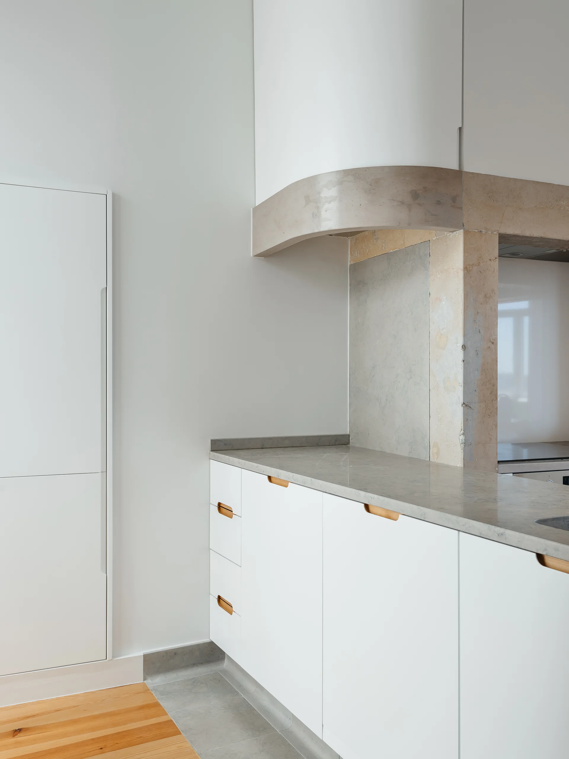A minimalist kitchen features white flat-front lower cabinets with wood recessed pulls, a grey stone countertop, and a curved stone range hood.