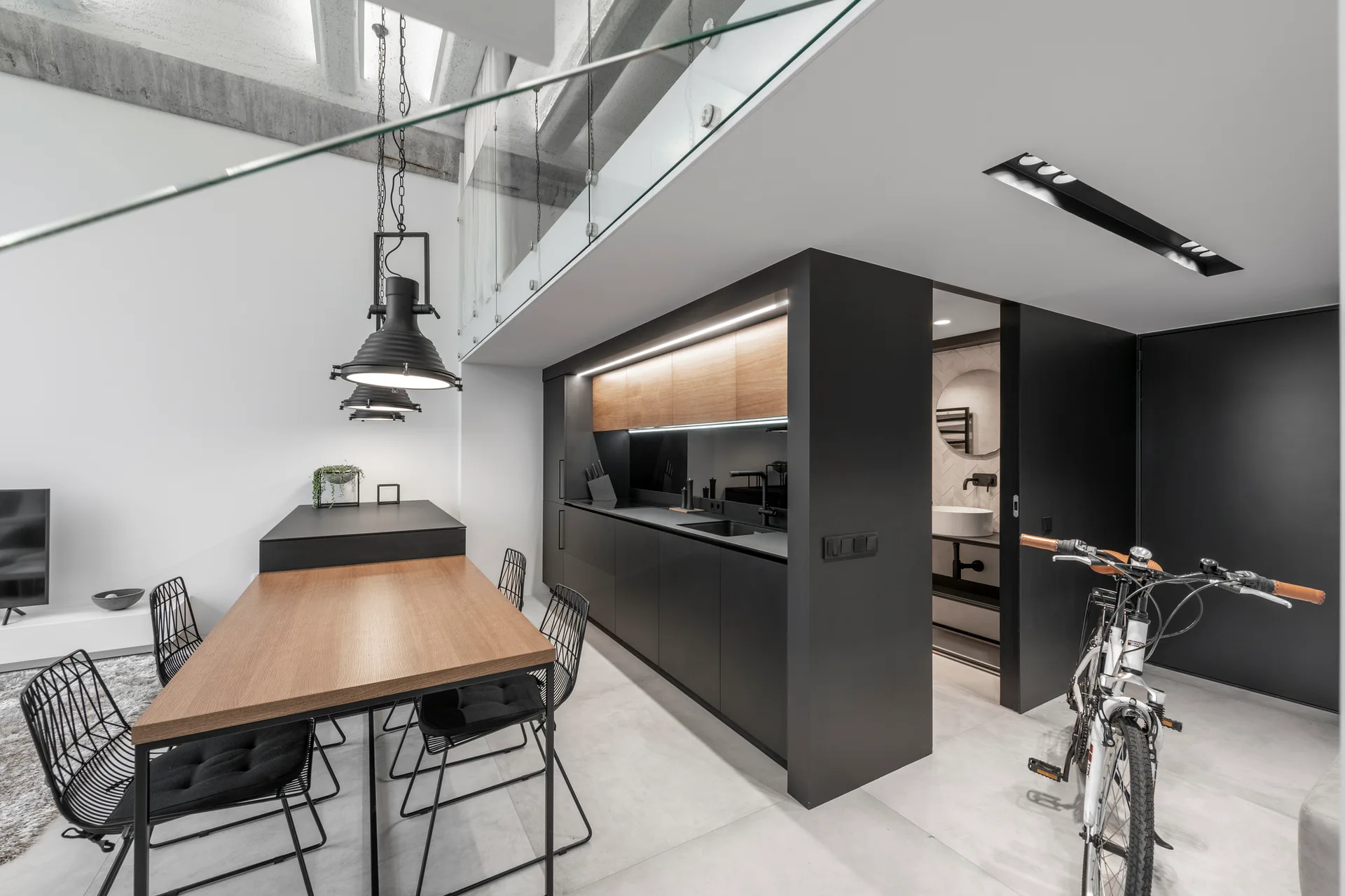 An industrial loft kitchen features matte black cabinetry, a wood dining table with wireframe chairs, and exposed concrete ceiling beams.