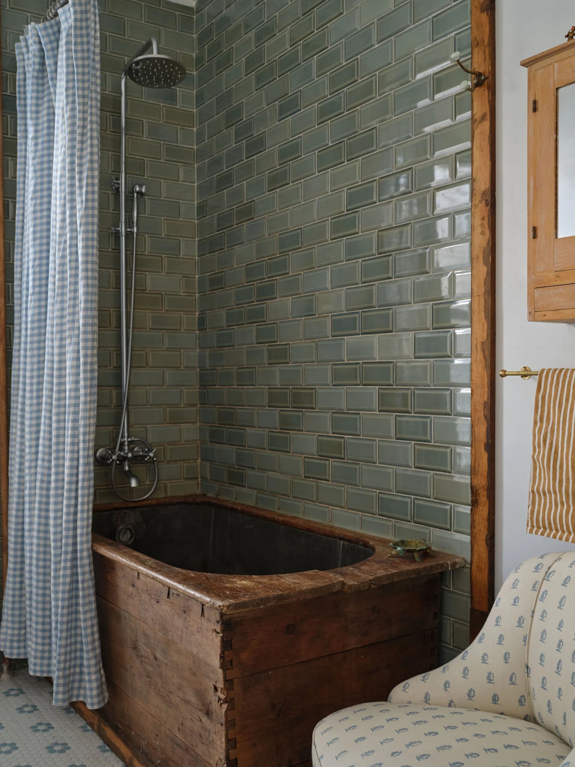 A rustic bathroom features a wooden box-frame bathtub beneath a chrome showerhead set against glossy sage green subway tiles and a blue gingham curtain.