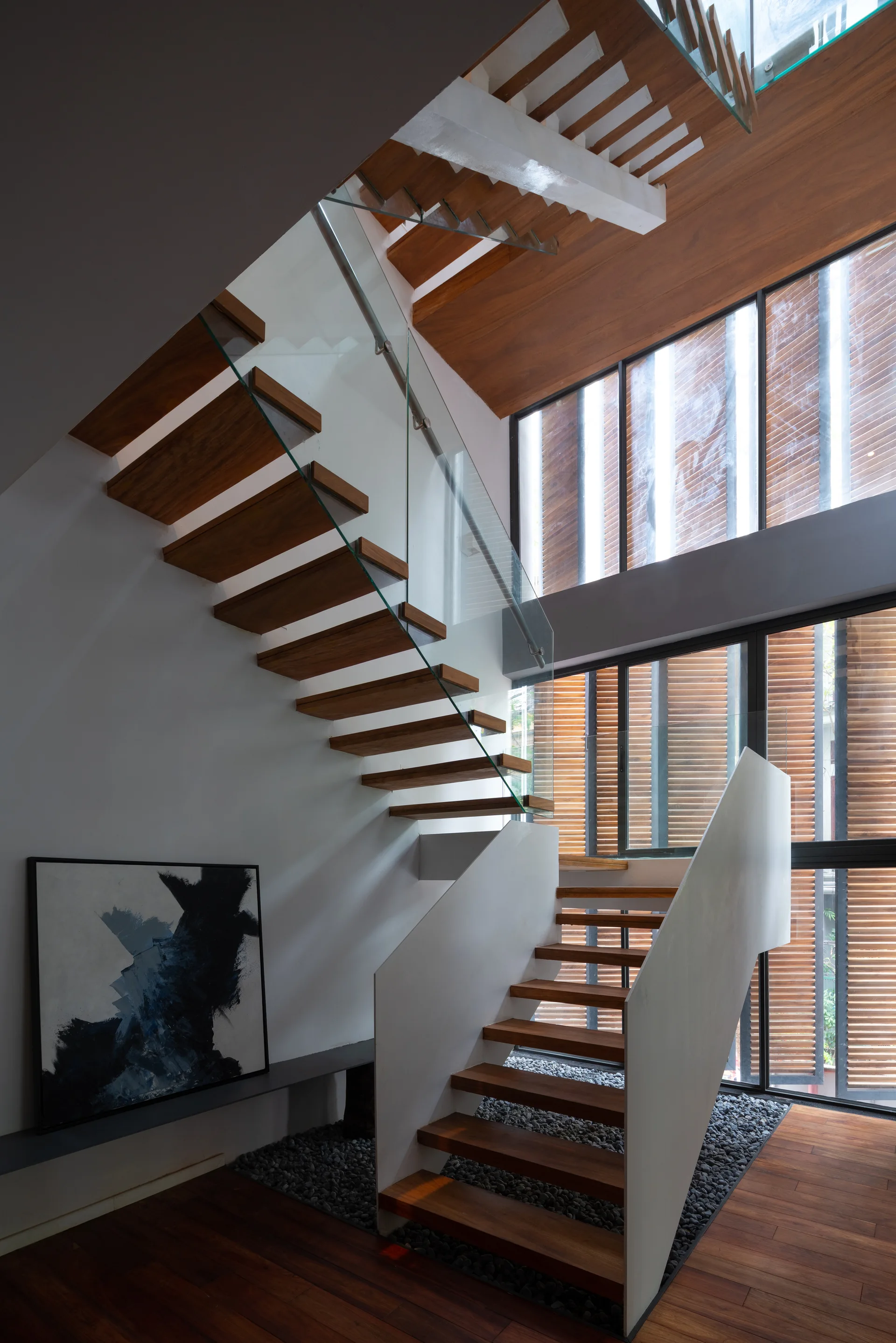 A modern floating wood staircase with a glass railing climbs past white walls and a tall window with slatted wood blinds, featuring a pebble bed at the base.