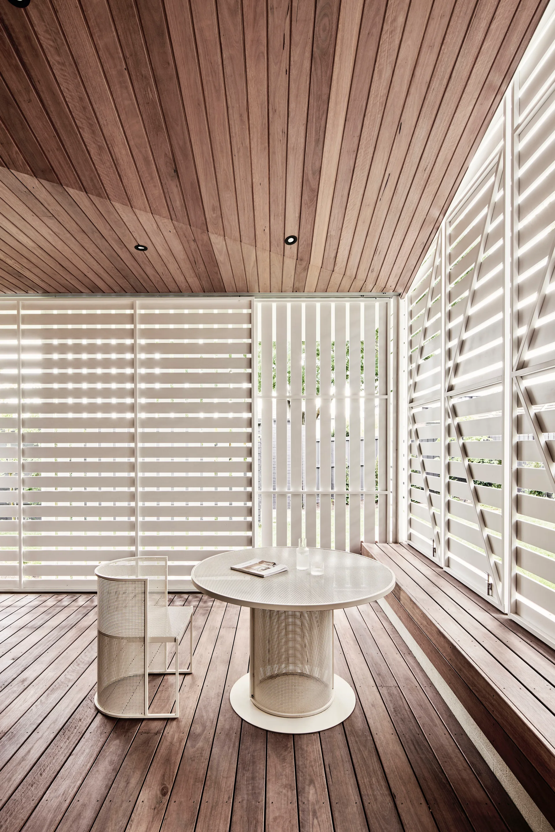 A modern covered patio with wood plank flooring, a matching wood ceiling, white slatted screens, and a white round table.