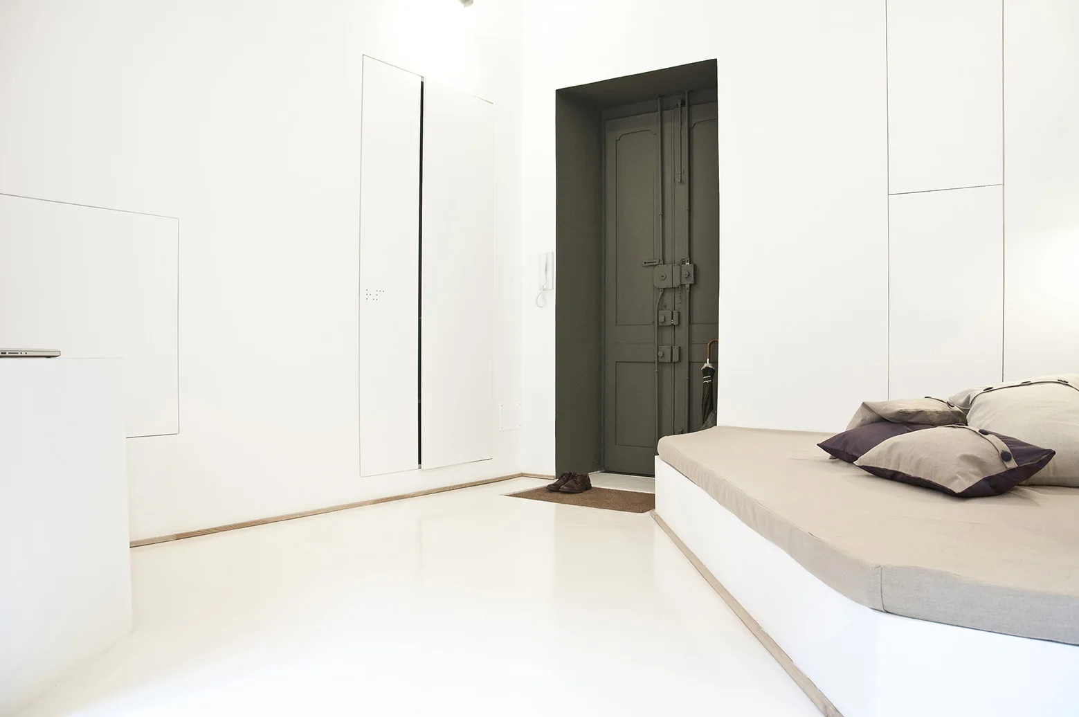 A minimalist white entryway featuring a recessed dark grey door, flush built-in cabinetry, glossy white floors, and a low platform daybed.