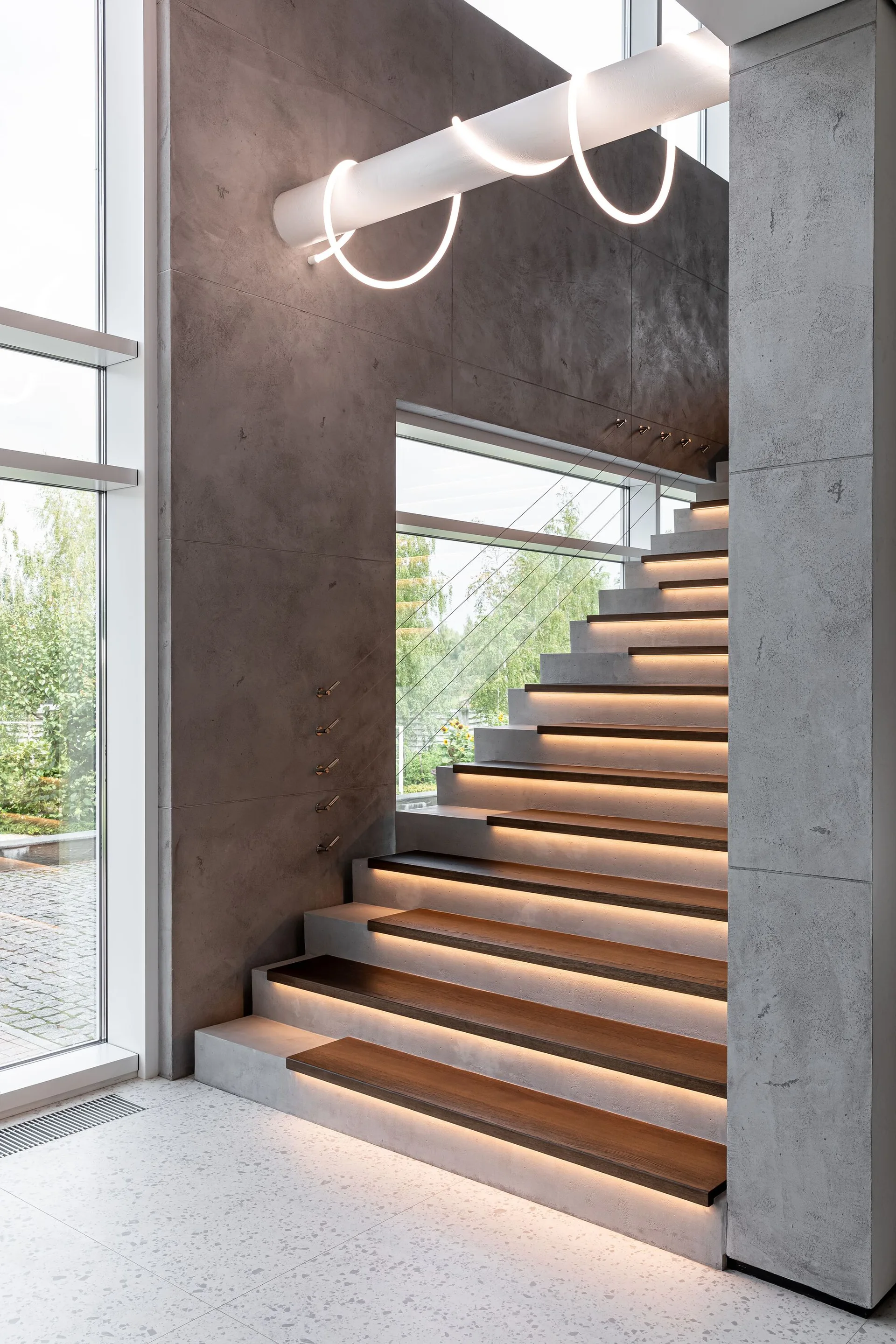 A modern concrete staircase features dark wood treads illuminated by warm under-step lighting, set against large gray wall panels and terrazzo flooring.