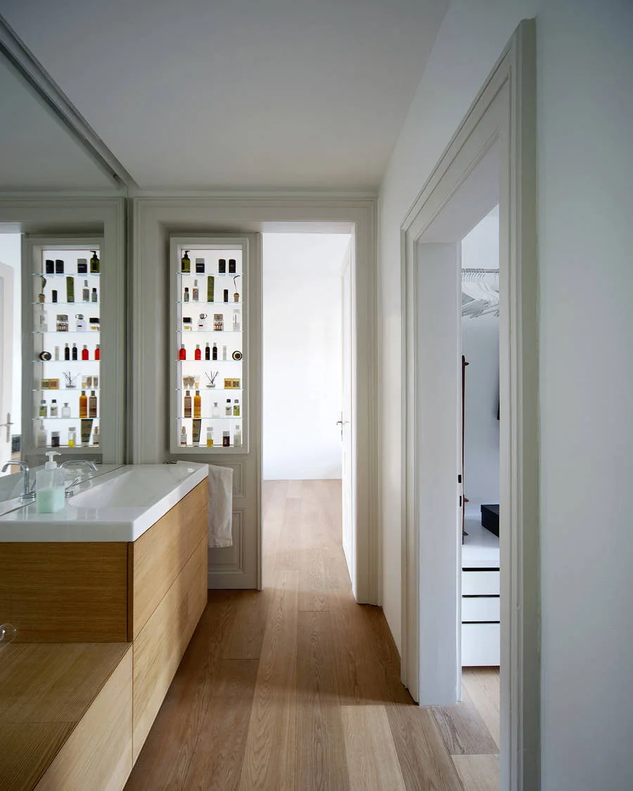 A modern bathroom with a light wood vanity, large mirror, and a narrow recessed wall niche with glass shelves displaying colorful toiletries.