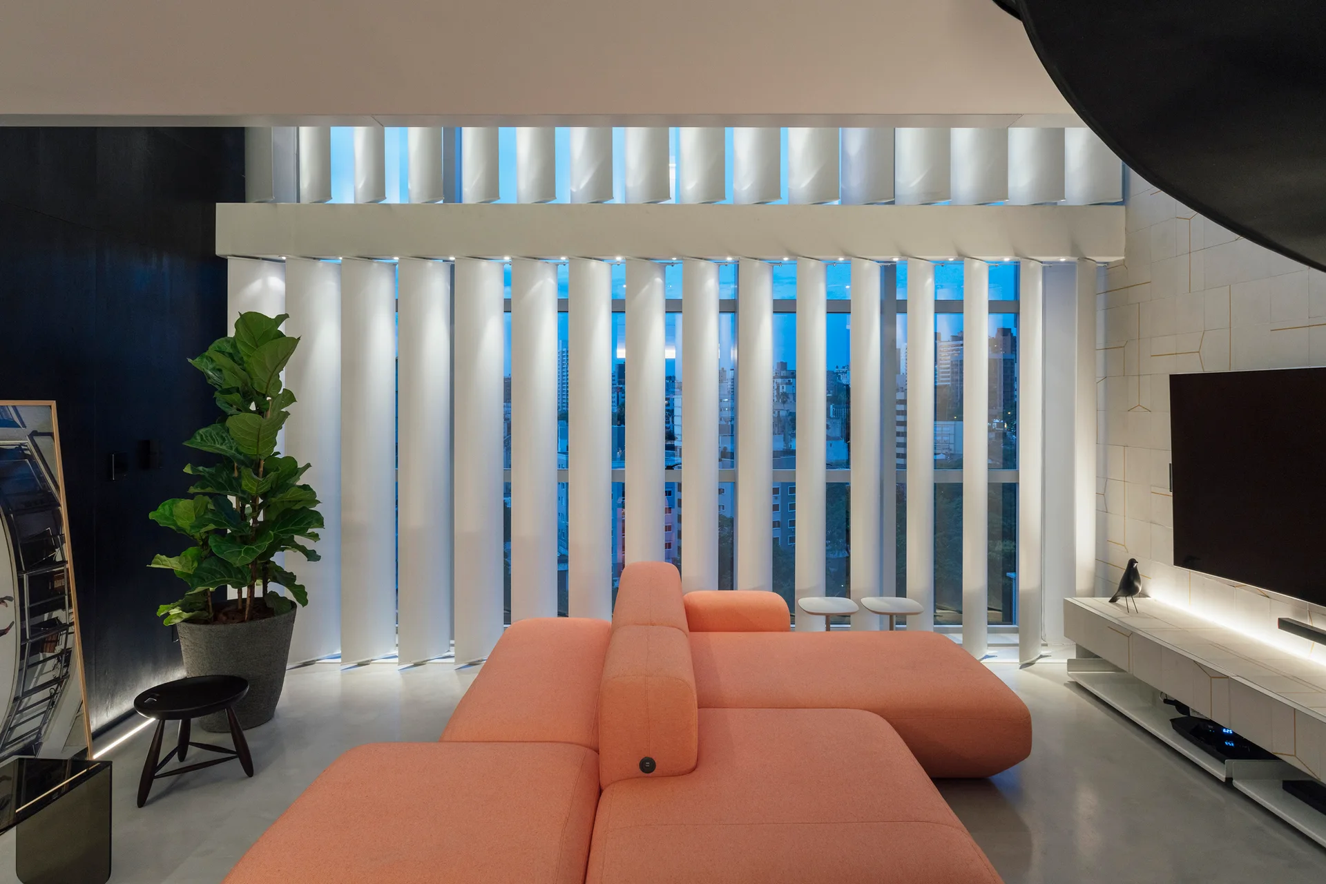 A modern living room featuring a large coral-colored sectional sofa, floor-to-ceiling windows with white vertical blinds, and a wall-mounted TV.