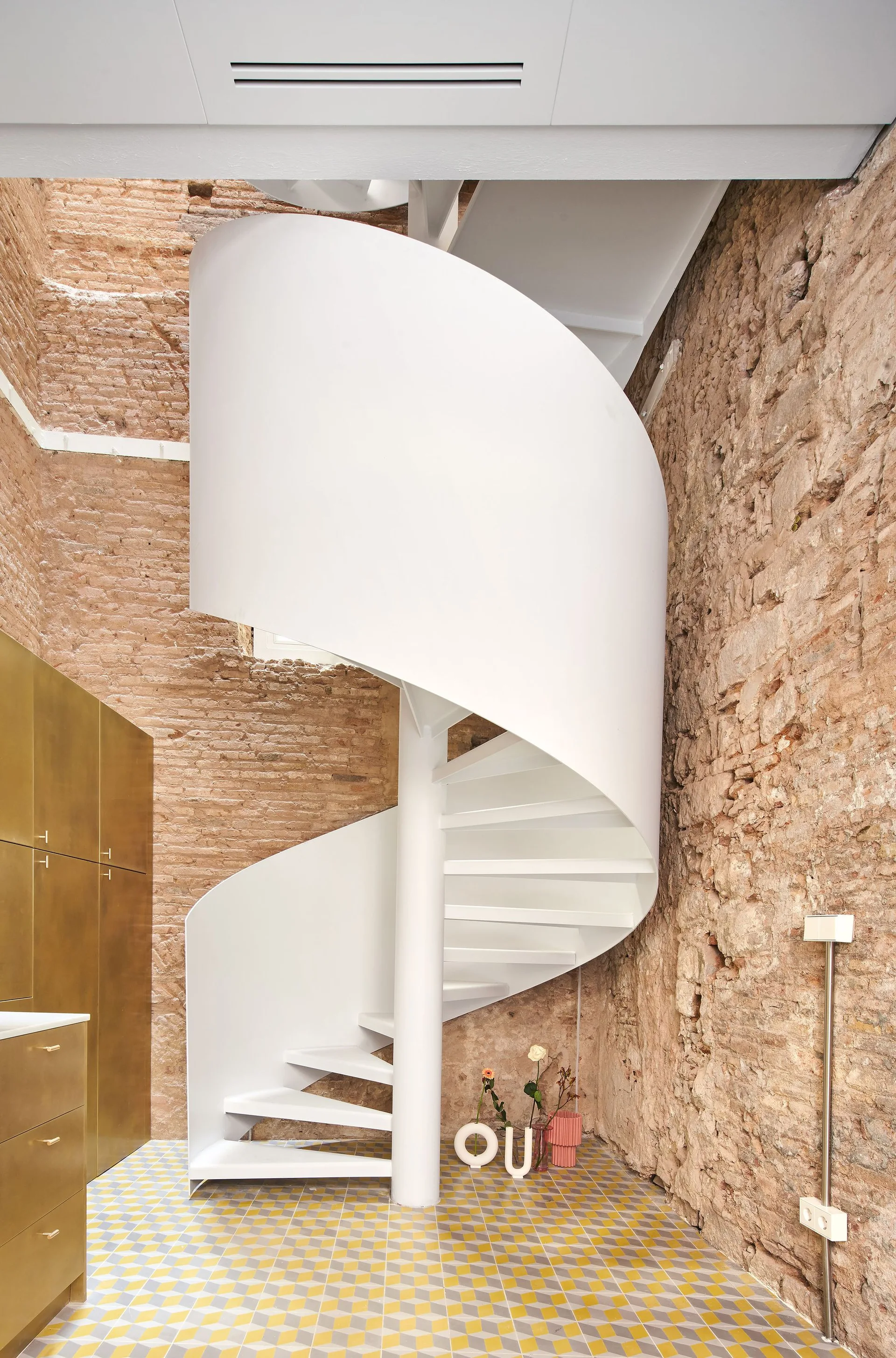 A white metal spiral staircase curves upward in front of a rustic exposed brick wall, with yellow geometric tile flooring below and brass cabinets nearby.