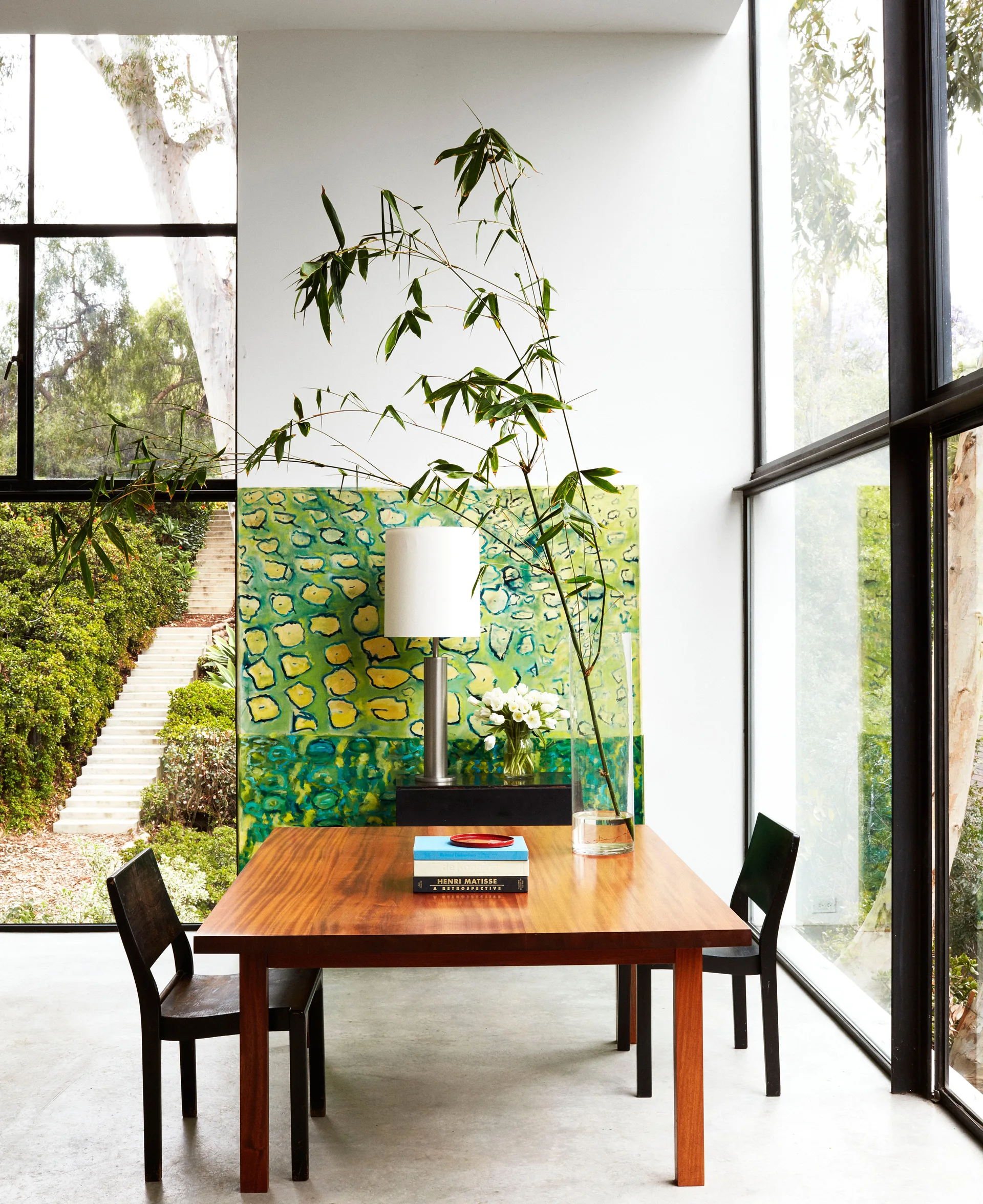 A minimalist dining room features a wooden table, black chairs, a tall glass vase with bamboo branches, and floor-to-ceiling windows looking outside.