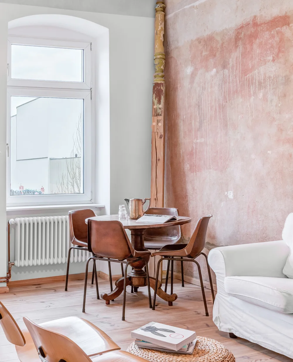 A round wooden dining table surrounded by brown leather chairs stands next to a distressed pink plaster wall and a white window.